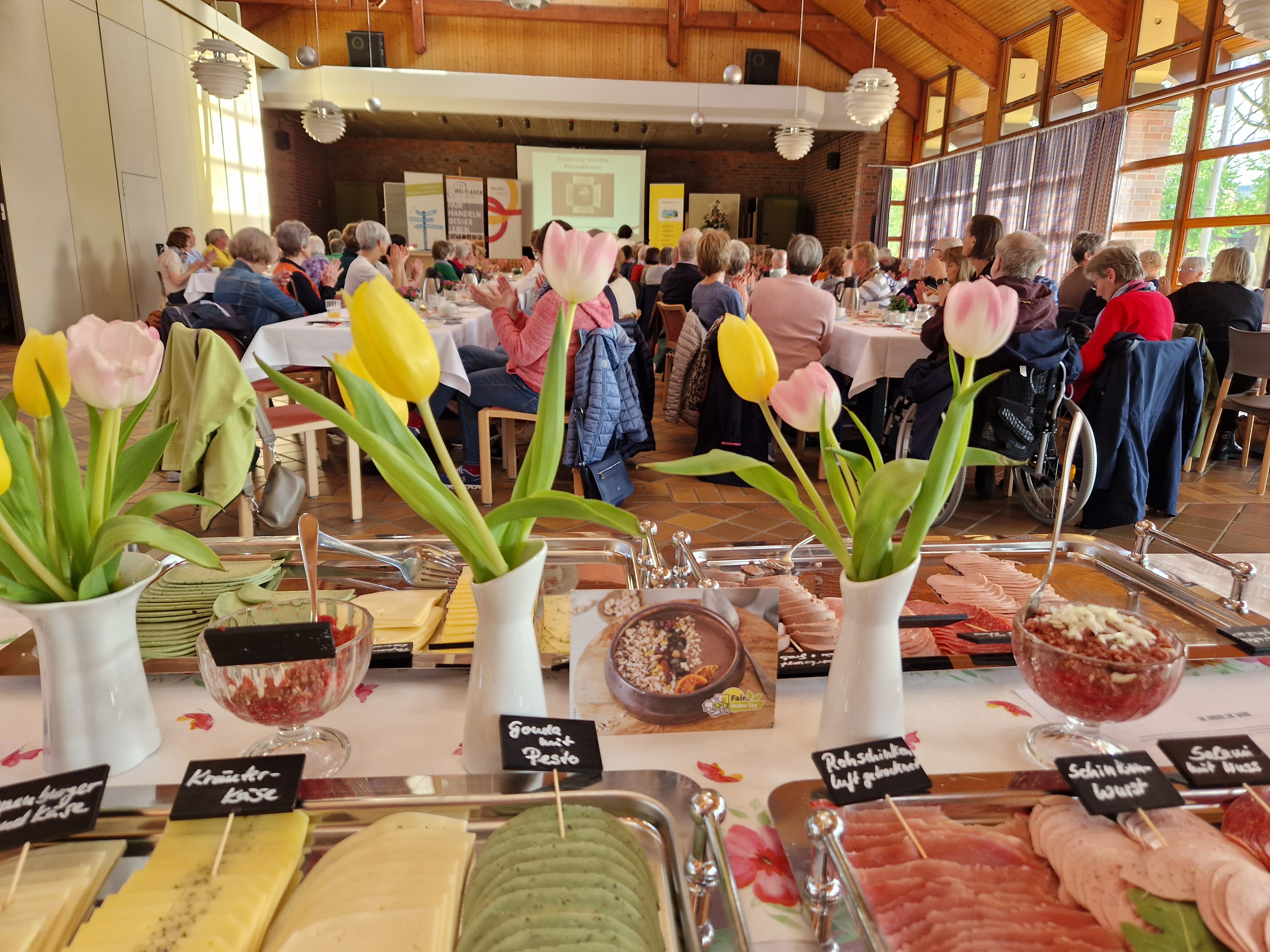 Ein Saal mit Tischen und Menschen beim Essen Im Vordergrund ein Buffet-Tisch mit Essen und gelben Tulpen