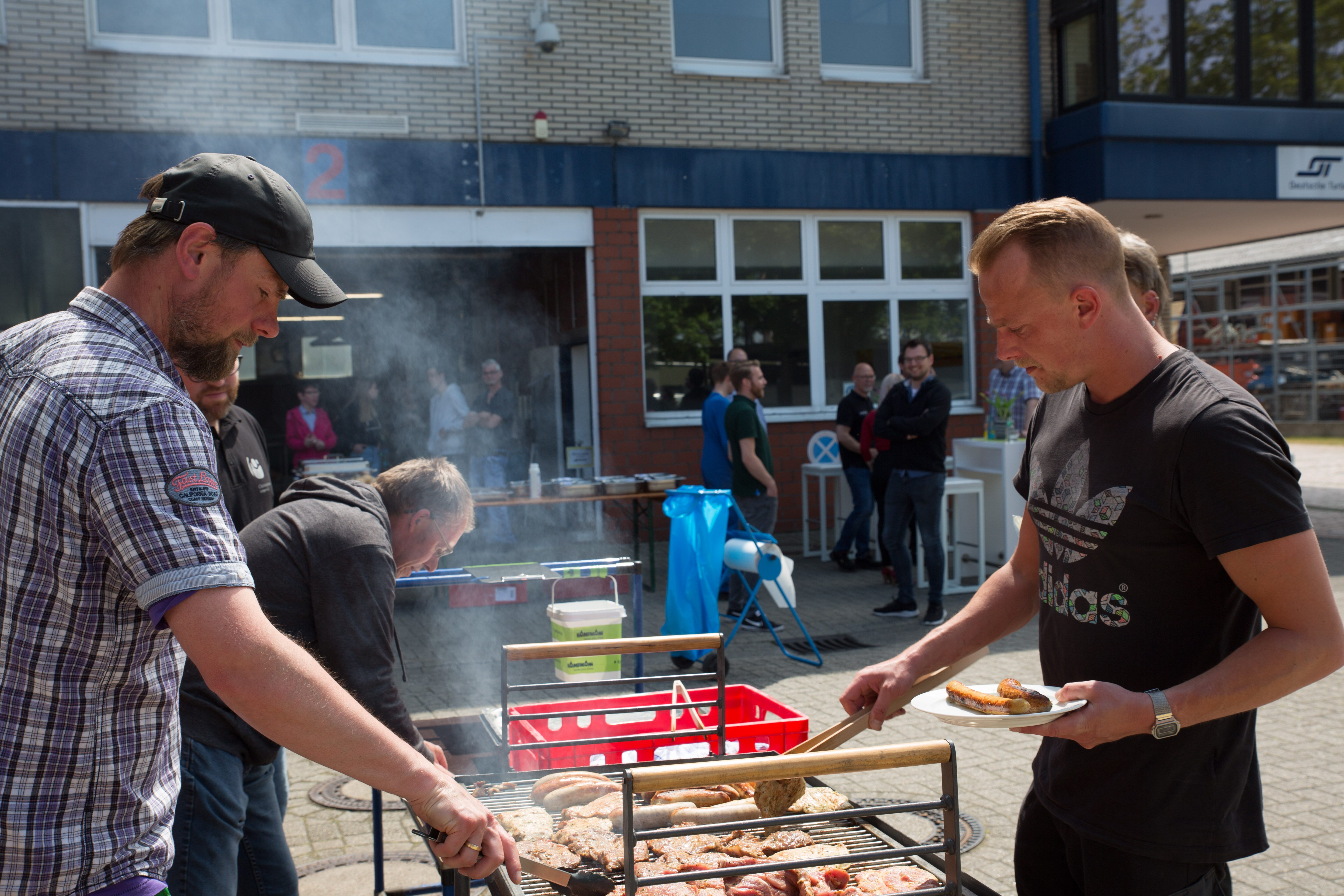 Menschen auf einem Fest. Im Vordergrund zwei Männer an einem Grill mit Würstchen und Steaks