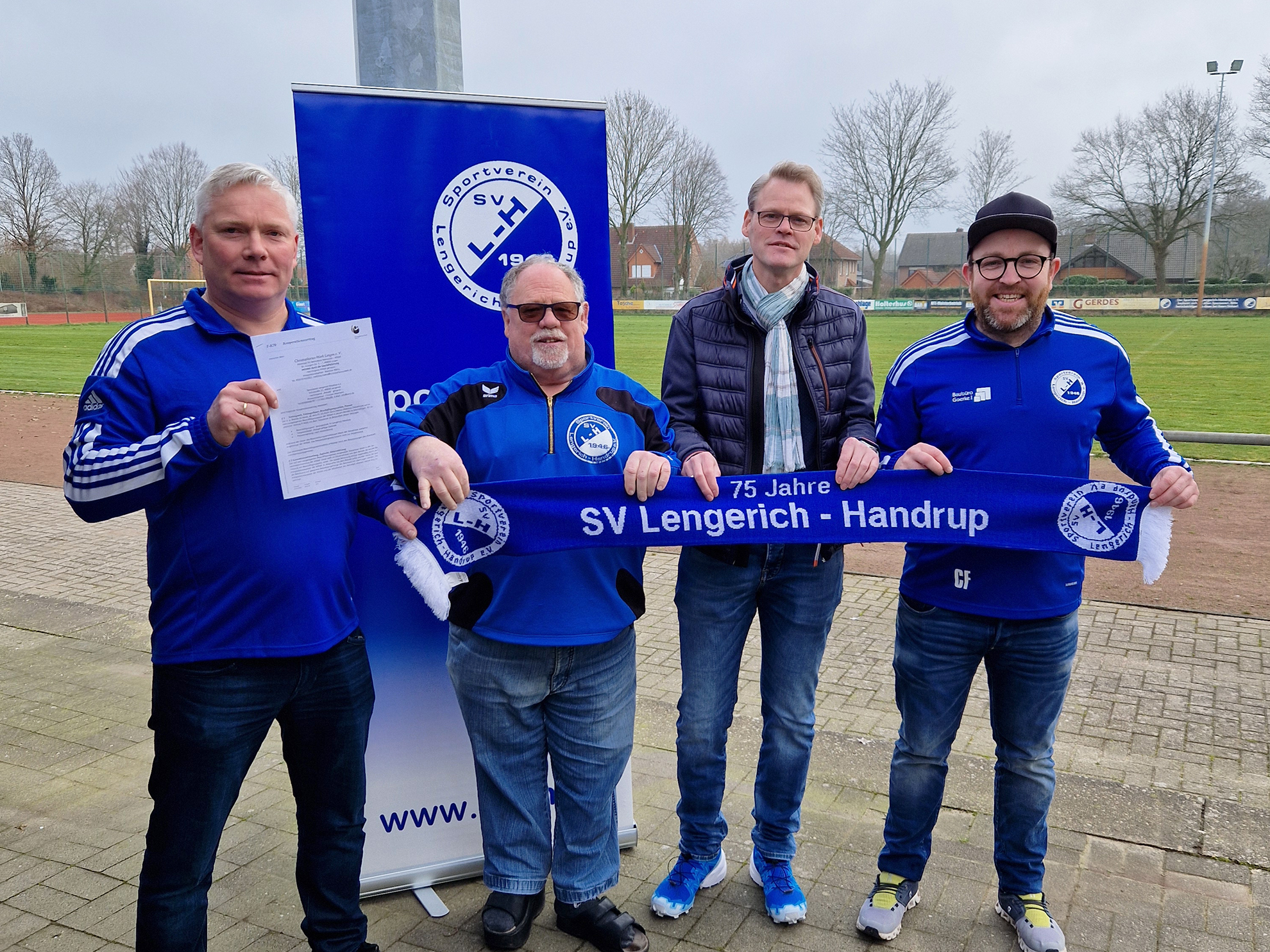 Man sieht vier Männer in blauen Sportjacken vor einem Sportplatz. Ein Mann hält ein Blatt Papier in den Händen, die anderen drei halten gemeinsam einen Vereinsschal.