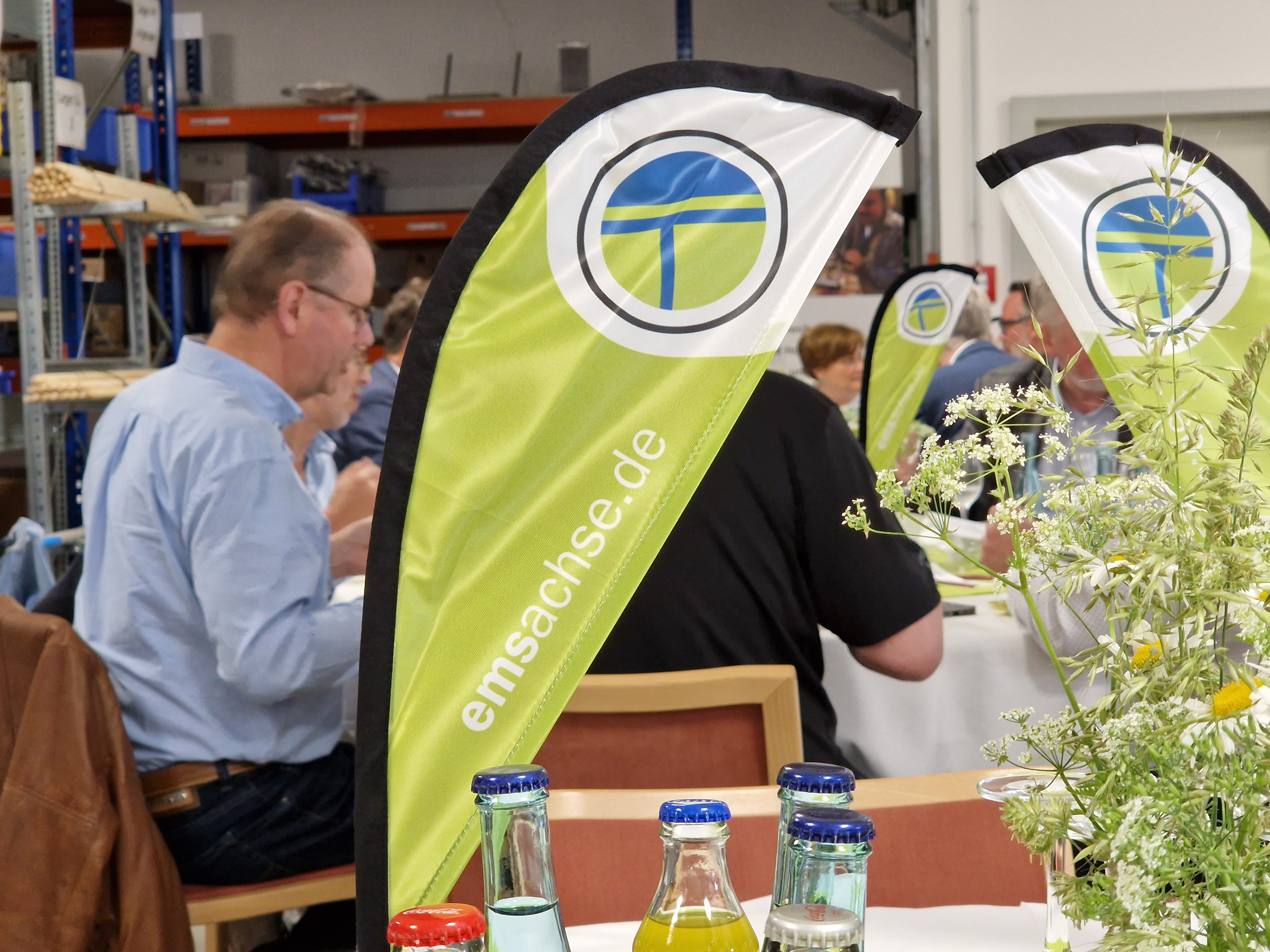 Im Hintergrund sitzen Menschen an Tischen und essen. Im Vordergrund steht ein grüner Tischaufsteller mit der Aufschrift emsachse.de