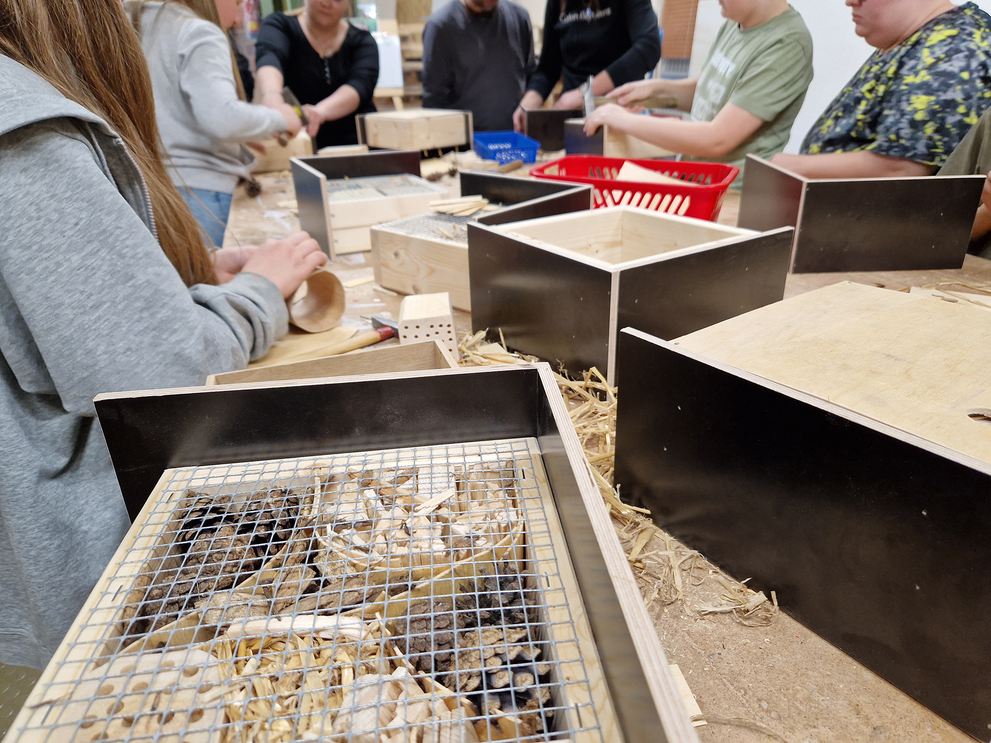 Das Bild zeigt eine Gruppe von Menschen, die gemeinsam an einem Tisch stehen und handwerkliche Arbeiten verrichten. Im Vordergrund sind mehrere Insektenhotels zu sehen, die teilweise bereits mit Naturmaterialien wie Holzstücken und Stroh befüllt sind. Einige Personen im Hintergrund bearbeiten Holzstücke und andere Materialien, um die Insektenhotels fertigzustellen.