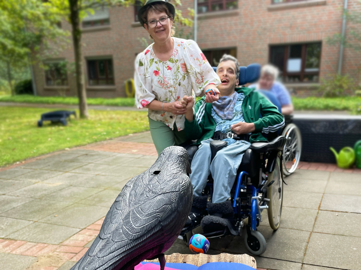 Eine Person im Rollstuhl wirft mit der Hilfe einer weiteren neben ihr stehenden Person einen Ball auf einen künstlichen Vogel.