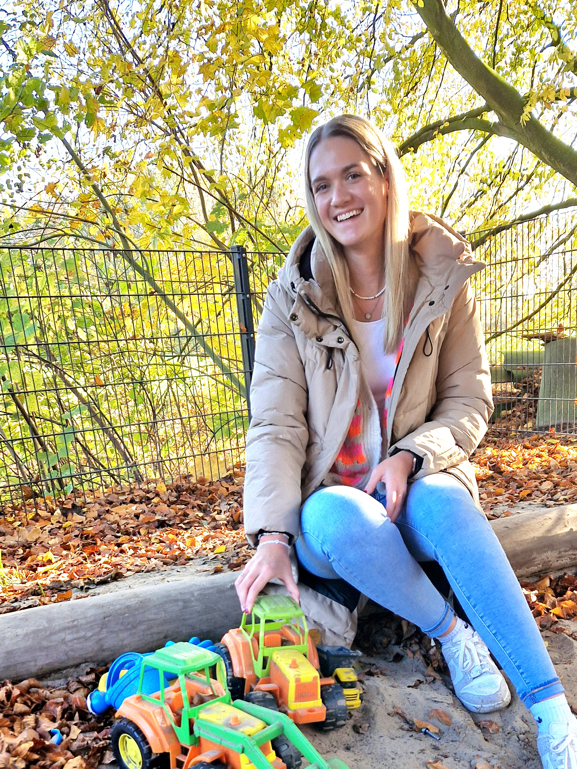 Eine junge Frau sitzt am Rand eines Sandkastens. Im Hintergrund leuchten herbstliche Bäume. 