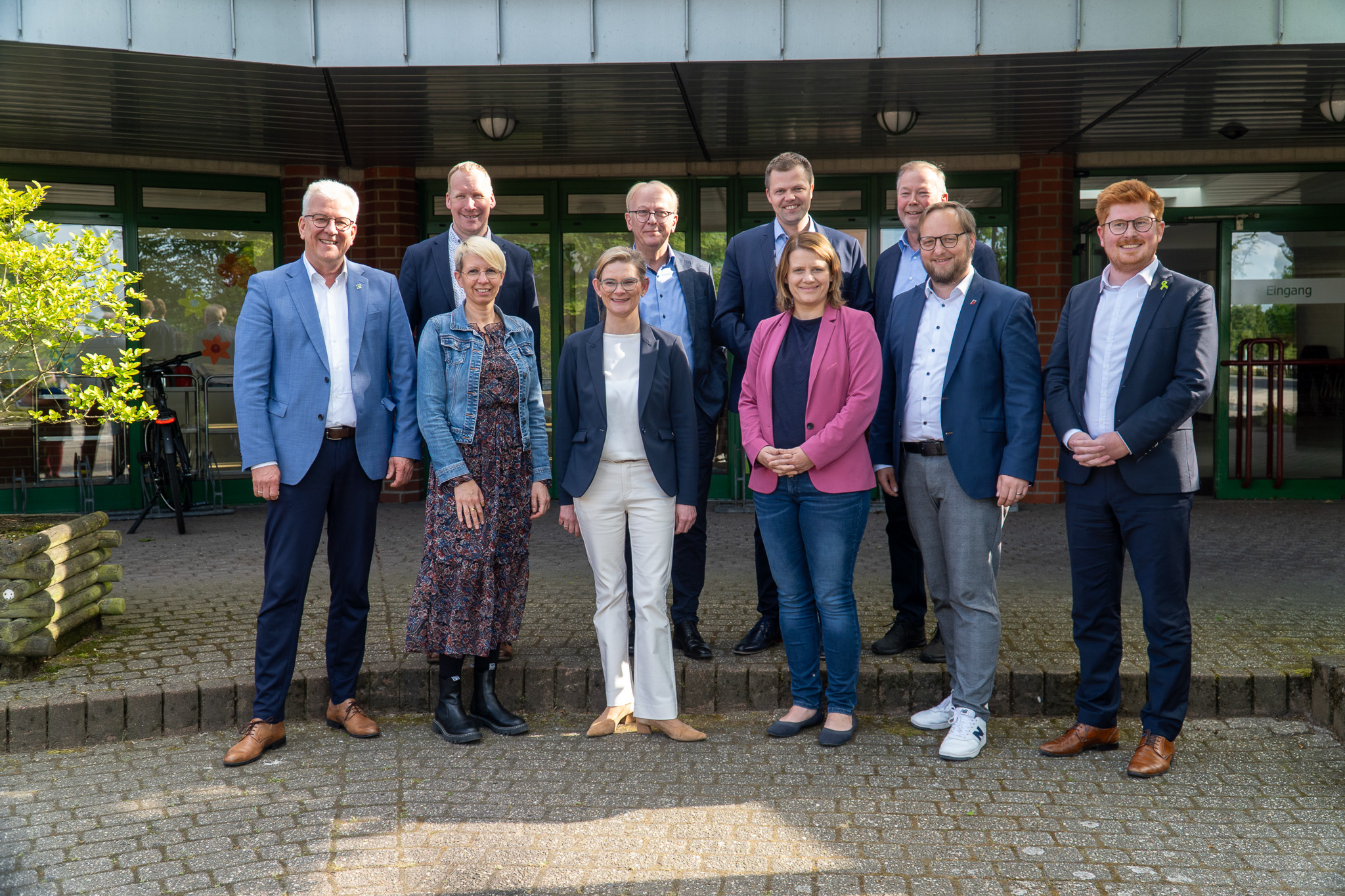 Vor dem Eingang der Jakob-Muth-Schule in Meppen steht eine Gruppe von zehn Personen für ein gemeinsames Foto. Alle blicken lächelnd in die Kamera. Die Gruppe besteht aus Vertreterinnen und Vertretern der drei emsländischen Träger der Behindertenhilfe, des Landkreises Emsland sowie aus Mitgliedern des niedersächsischen Landtags.  Im Vordergrund stehen sechs Personen nebeneinander, darunter drei Frauen in Blazern, eine Frau in einem geblümten Kleid mit Jeansjacke sowie drei Männer in Anzug oder Sakko. Hinter ihnen stehen fünf weitere Männer, ebenfalls formell gekleidet.  Im Hintergrund ist der Eingangsbereich der Schule zu sehen: ein überdachtes Vordach mit Glas- und Klinkerelementen sowie eine grüne Tür mit dem Schild „Eingang“. Links im Bild ist ein Baum mit frischem Grün zu erkennen. Die Stimmung ist freundlich und offen.