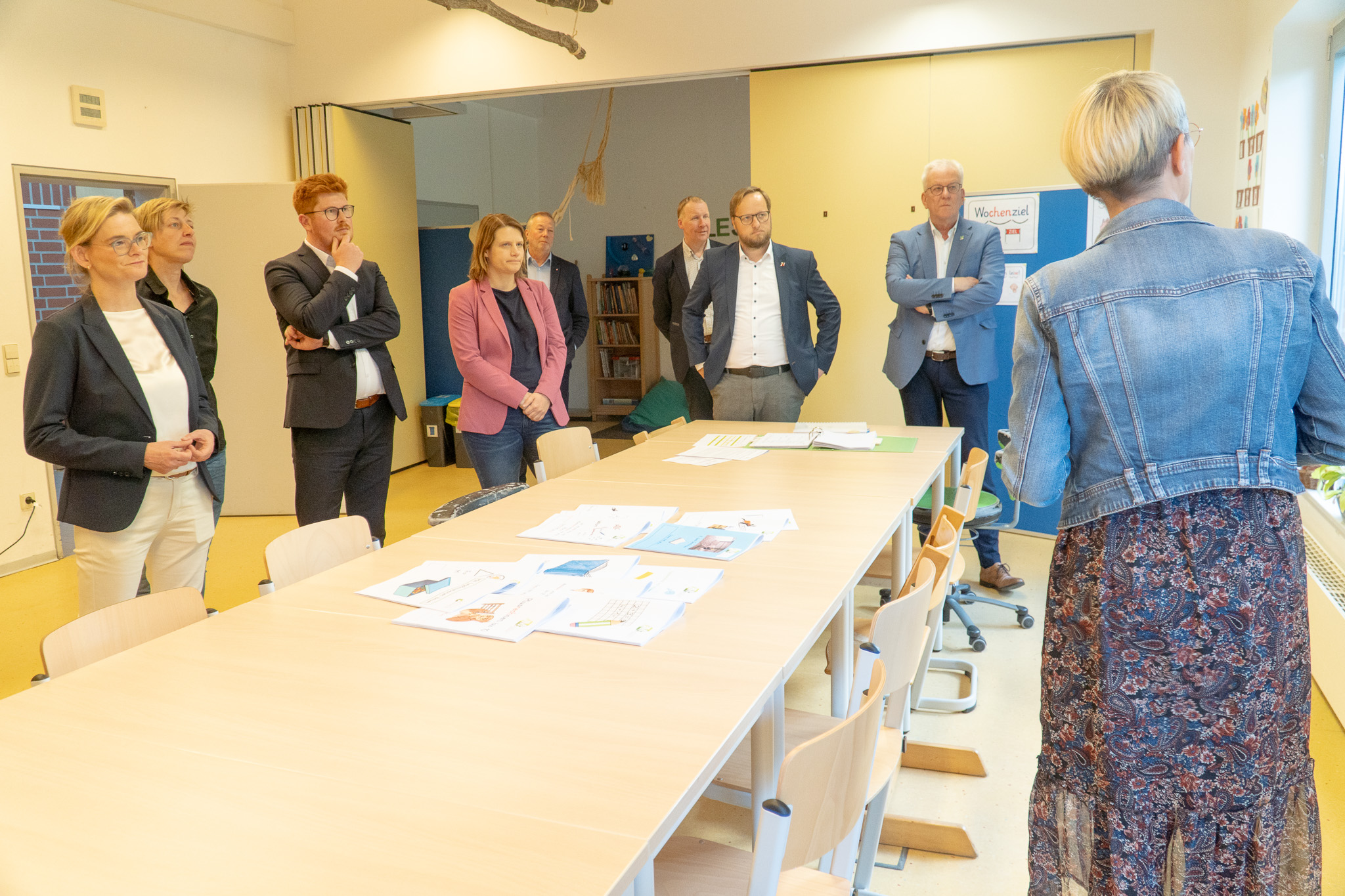 In einem hellen Klassenraum der Jakob-Muth-Schule steht eine Gruppe von neun Erwachsenen um einen rechteckigen Tisch. Die meisten Personen stehen an der gegenüberliegenden Seite des Tisches und hören aufmerksam einer Frau im Vordergrund zu, die mit dem Rücken zur Kamera spricht. Sie trägt ein geblümtes Kleid und eine Jeansjacke. Auf dem Tisch liegen zahlreiche bunte Unterrichtsmaterialien, Arbeitsblätter und Ordner ausgebreitet.  Die Zuhörenden stehen locker, aber aufmerksam. Einige haben die Hände verschränkt oder in den Hosentaschen. Im Hintergrund ist eine Regalwand mit Büchern und Kissen zu sehen, an der rechten Wand ein blaues Whiteboard mit der Aufschrift „Wochenziel“. Die Atmosphäre wirkt konzentriert und interessiert.