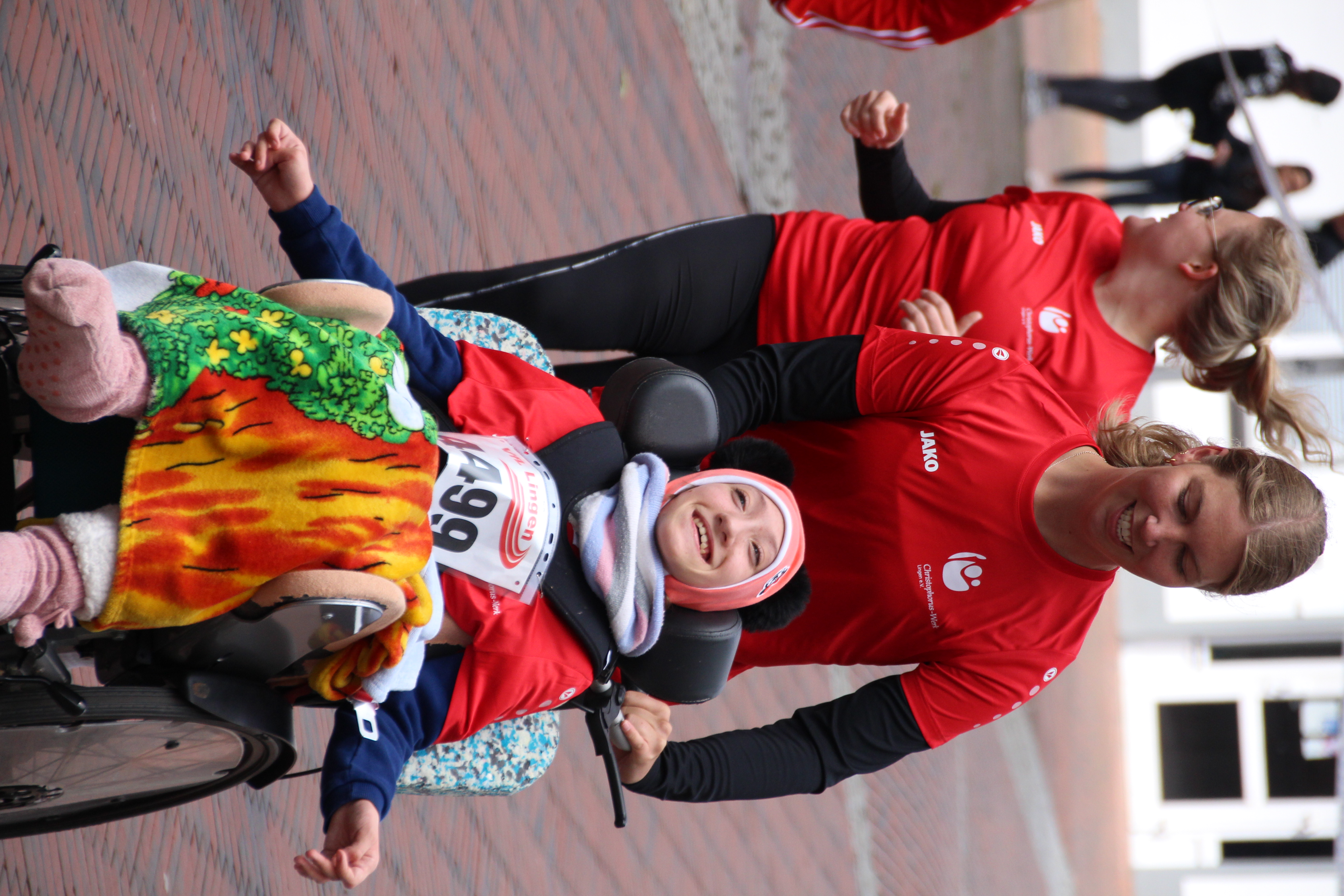 Eine junge Teilnehmerin im Rollstuhl lacht strahlend, während sie mit einem roten Trikot des Christophorus-Werks von einer Mitarbeiterin durch die Laufstrecke geschoben wird. Beide tragen Startnummern. Im Hintergrund laufen weitere Teilnehmende mit.