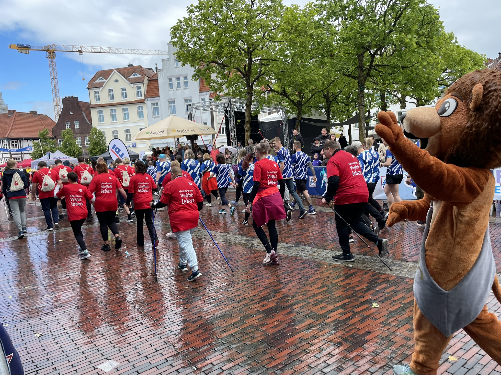 Eine große Gruppe von Teilnehmenden mit roten Laufshirts des Christophorus-Werks („Gemeinsam Vielfalt leben“) bewegt sich gemeinsam durch die Lingener Innenstadt. Am Bildrand winkt ein Maskottchen in Löwenkostüm. Die Stimmung ist fröhlich und bunt – trotz regennasser Straße.