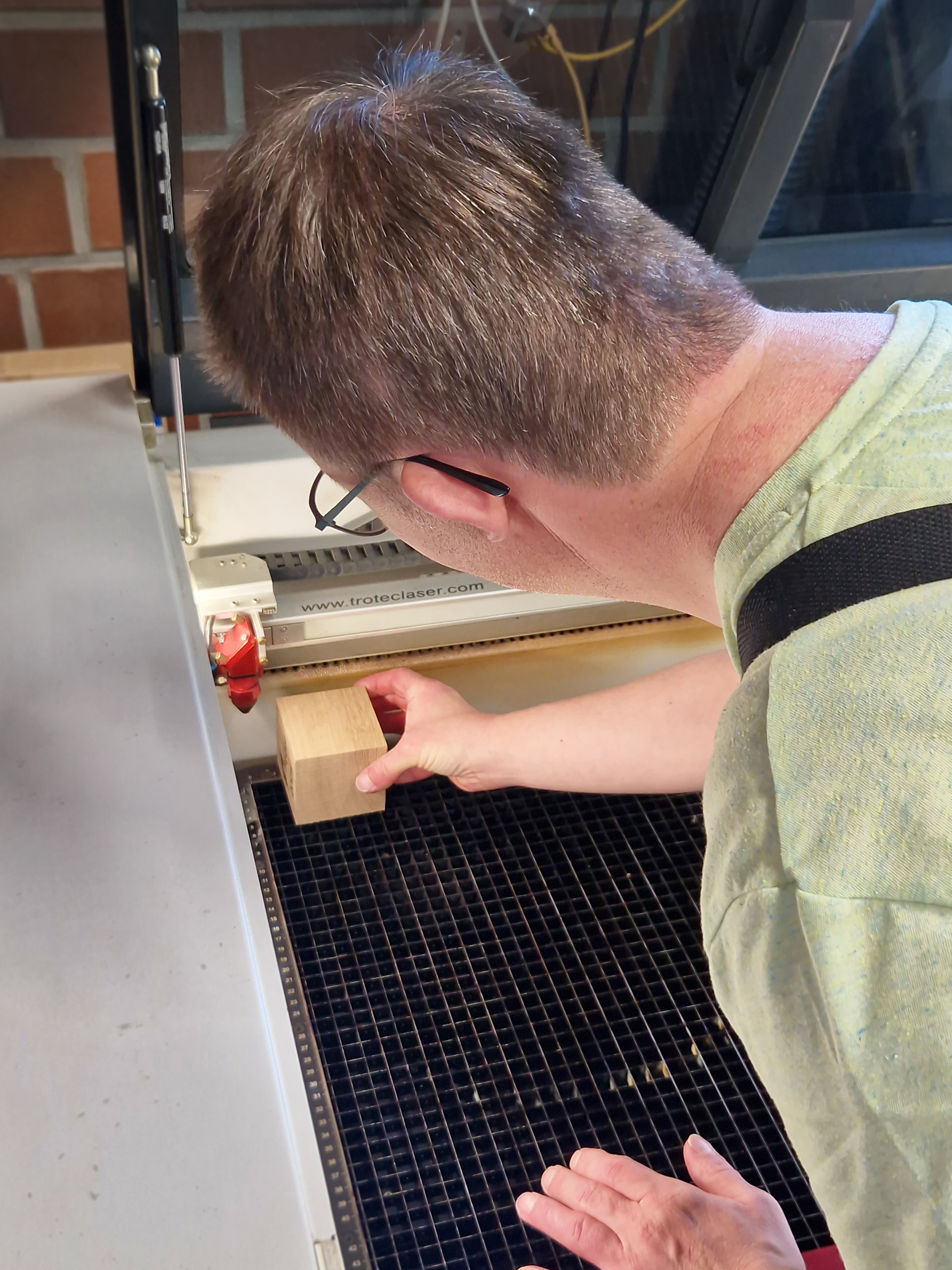 Ein Mann arbeitet mit einer Lasermaschine in der Werkstatt. Er legt den Holzwürfel in das Ge-rät ein, um die Gravur für den Pokal „Lingener Feder“ vorzubereiten.