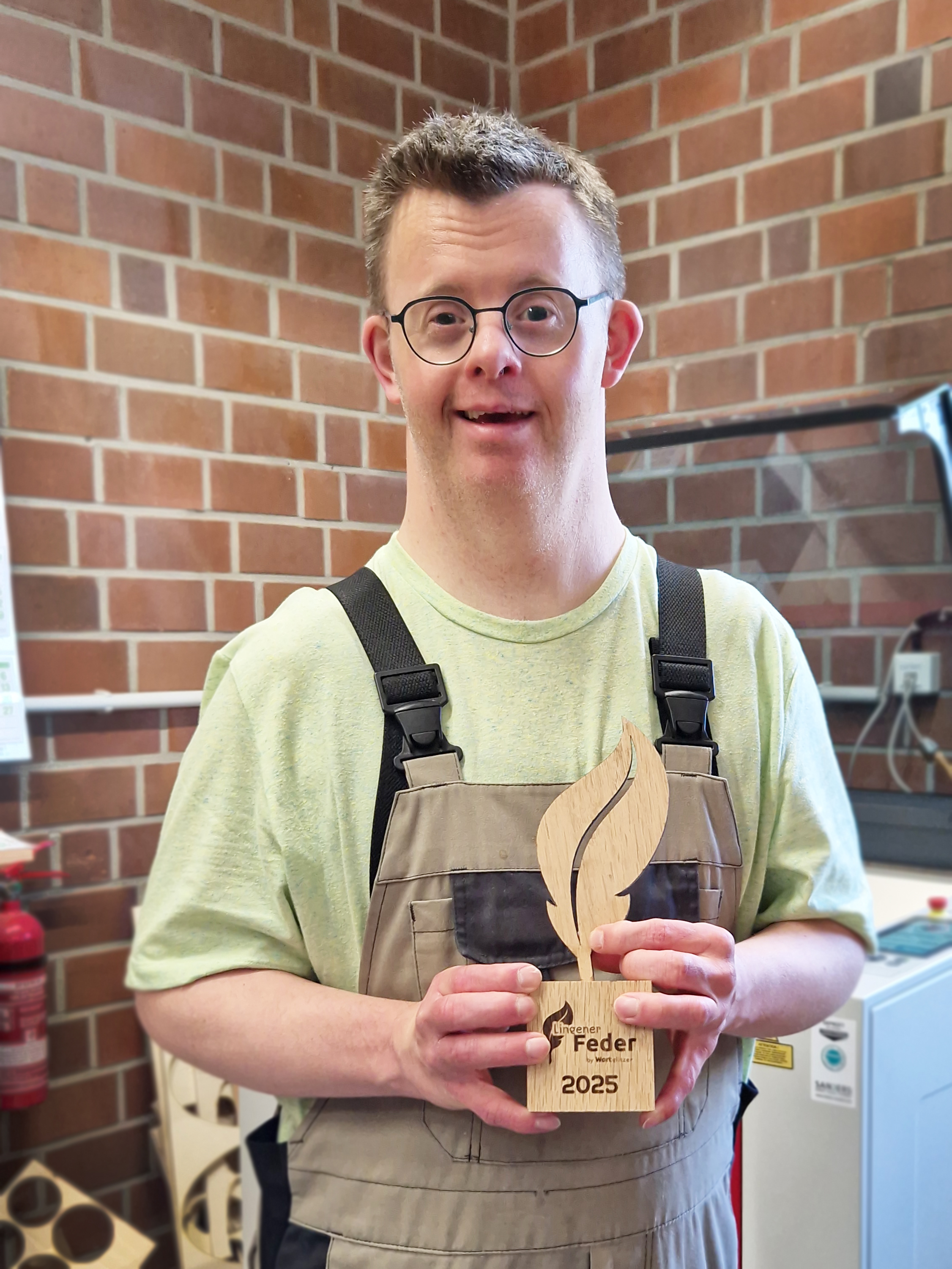 Ein Mann steht in der Holzwerkstatt und hält stolz den fertigen Pokal „Lingener Feder 2025“ in den Händen. Er lächelt in die Kamera, im Hintergrund ist eine Ziegelwand zu sehen.