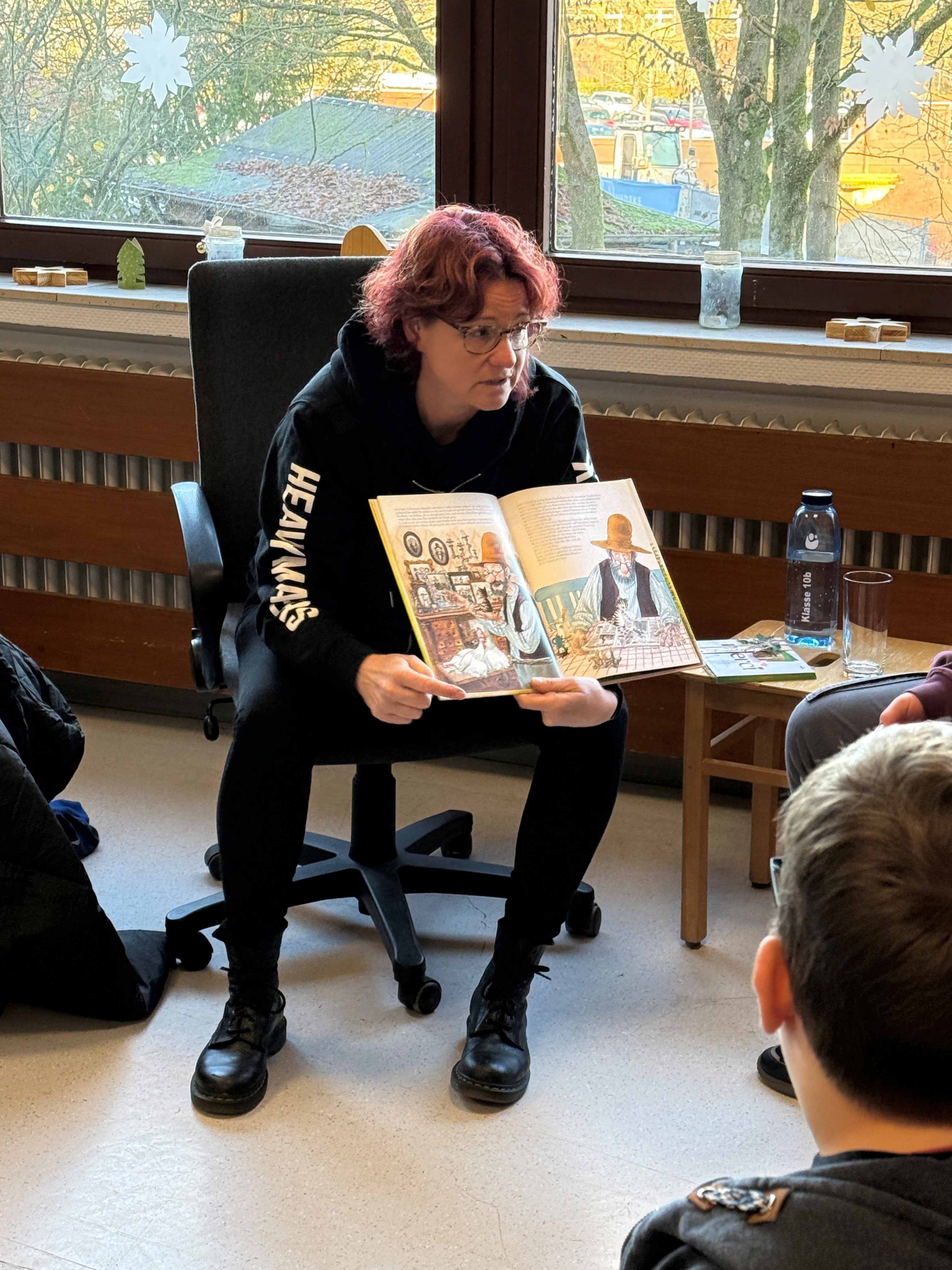 Eine Frau sitzt vor einem Fenster und h&auml;lt ein Buch aufgeschlagen in den H&auml;nden, aus dem sie ihrem Publikum etwas erkl&auml;rt.