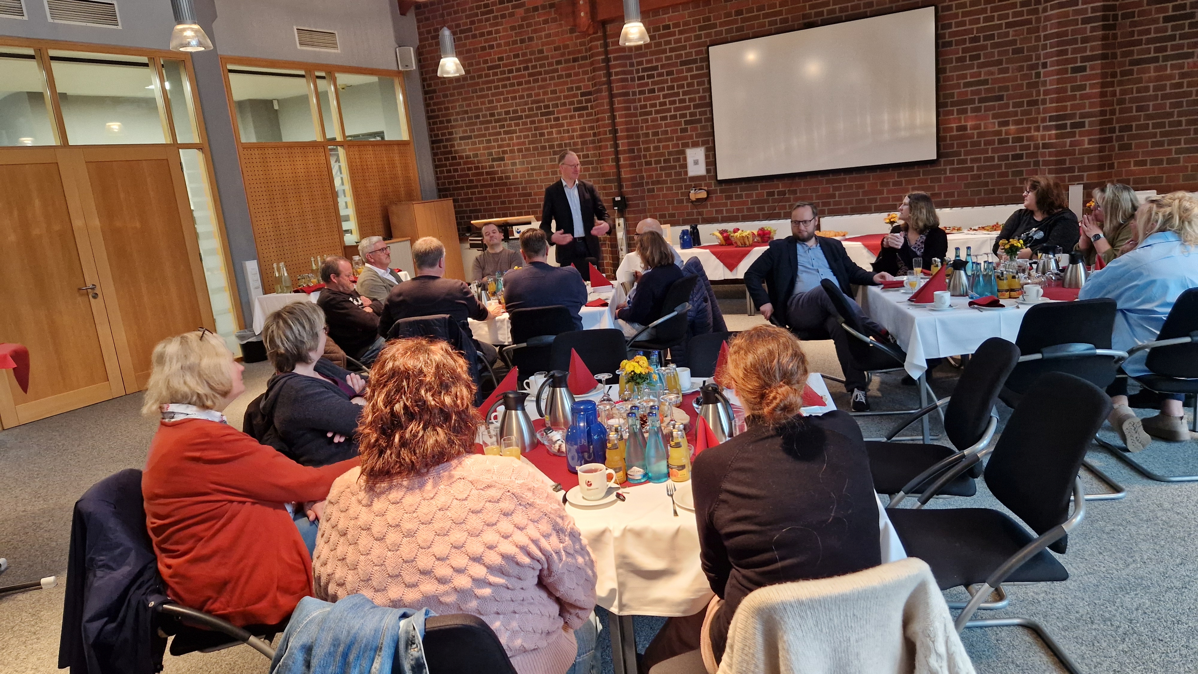 Hintergrund steht ein Mann, der eine Ansprache vor einer an eingedeckten Tischen sitzenden Personengruppe hält.