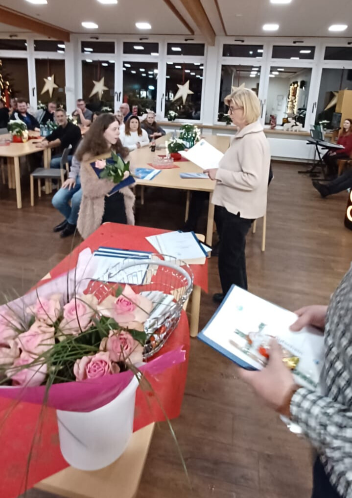 Ein gro&szlig;er Saal, auf einem Tisch stehen Rosen in einem Topf. Eine Frau h&auml;ndigt einer jungen Frau Blumen und ein Abschlusszeugnis &uuml;ber. 