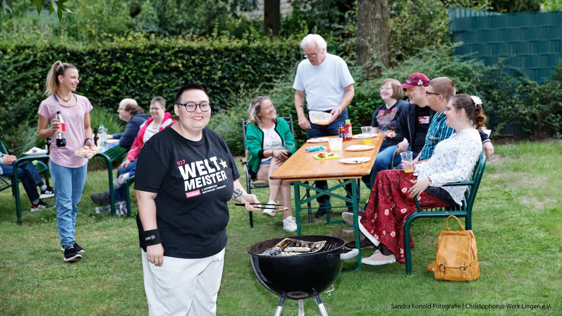 Eine Gruppe von Menschen mit und ohne Behinderung sitzt gemeinsam im Garten an Tischen. Im Vordergrund steht eine Person am Grill, im Hintergrund wird gelacht, gegessen und miteinander Zeit verbracht.