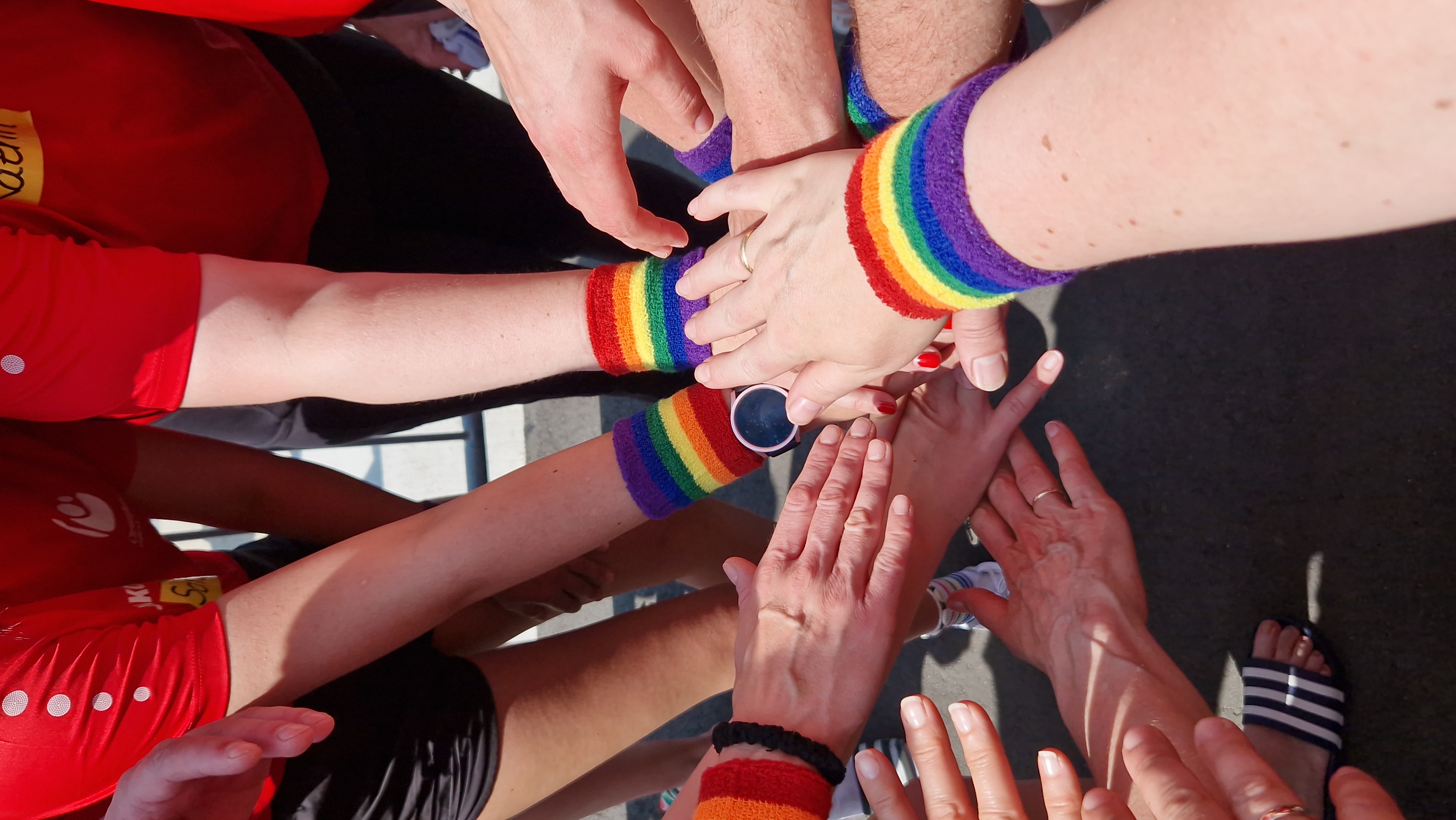 Nahaufnahme von zusammengelegten Händen der Teammitglieder mit bunten Regenbogen-Schweißbändern – ein Symbol für Teamgeist, Vielfalt und Zusammenhalt.