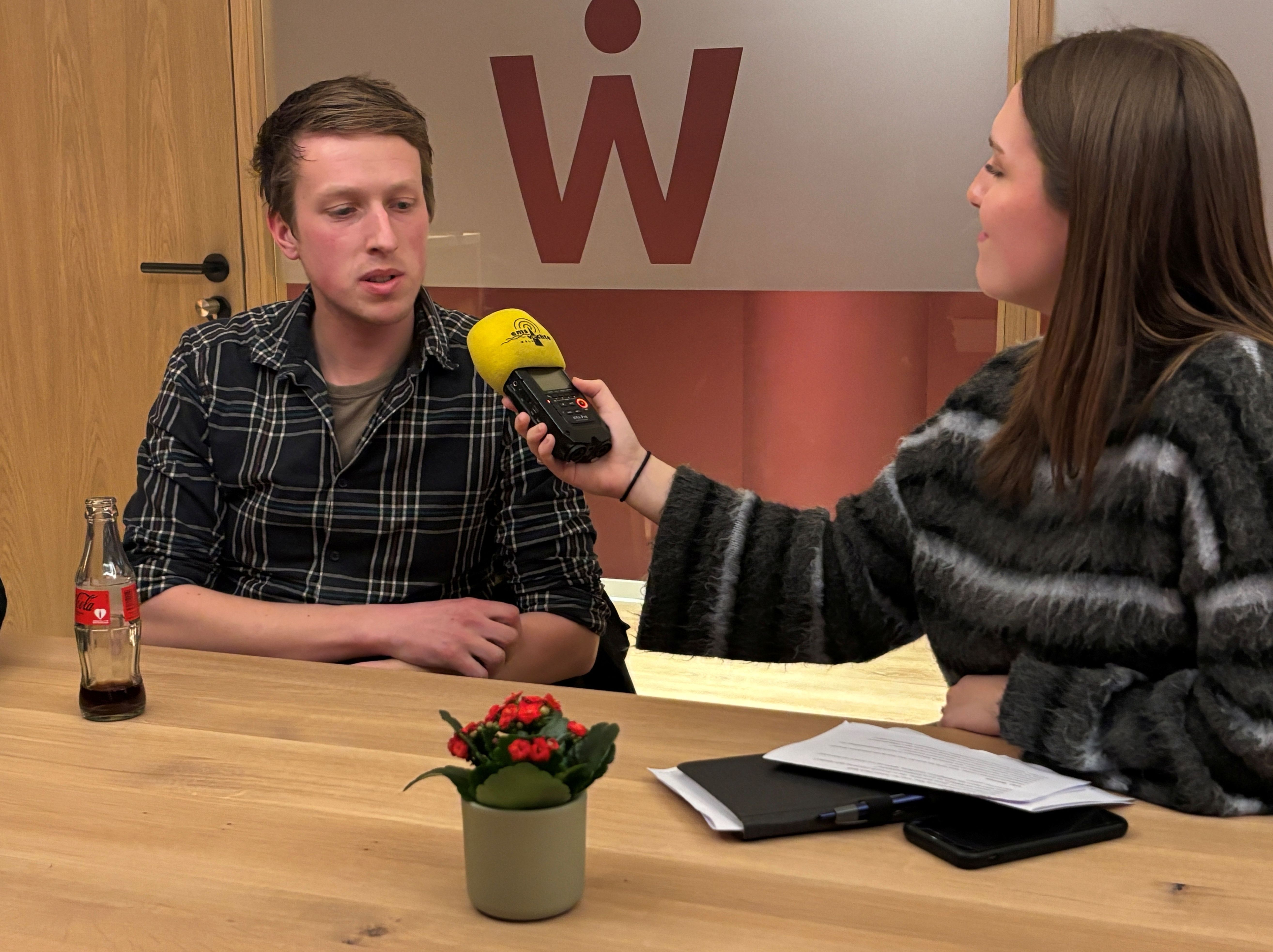 Eine Frau f&uuml;hrt ein Interview mit einem jungen Mann an einem Holztisch. Sie h&auml;lt ihm ein Aufnahmeger&auml;t mit gelbem Mikrofon entgegen. Der Mann sitzt ihr gegen&uuml;ber und spricht, vor ihm steht eine Glasflasche mit Getr&auml;nk. Auf dem Tisch liegen Notizen, ein Stift und ein Smartphone. Im Hintergrund ist eine Glaswand mit einem gro&szlig;en roten &bdquo;W&ldquo; zu sehen.