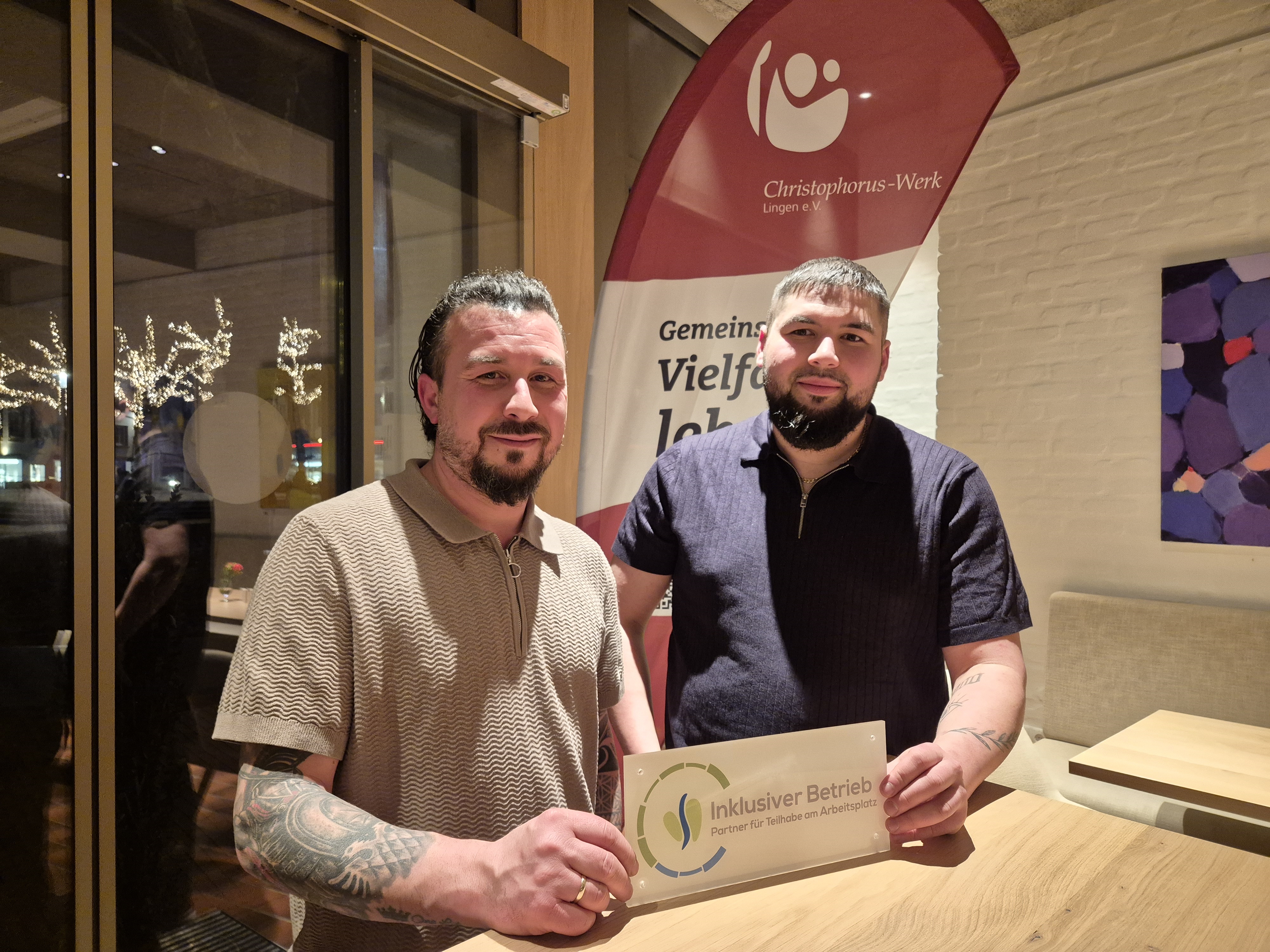 Zwei M&auml;nner stehen in einem Innenraum vor einem Roll-up-Banner des Christophorus-Werks Lingen e. V. und halten gemeinsam eine Plakette mit der Aufschrift &bdquo;Inklusiver Betrieb &ndash; Partner f&uuml;r Teilhabe am Arbeitsplatz&ldquo; in die Kamera. Sie stehen an einem Holztisch, l&auml;cheln und pr&auml;sentieren die Auszeichnung. Im Hintergrund sind ein Fenster mit Lichterdekoration sowie ein Wandbild zu sehen.