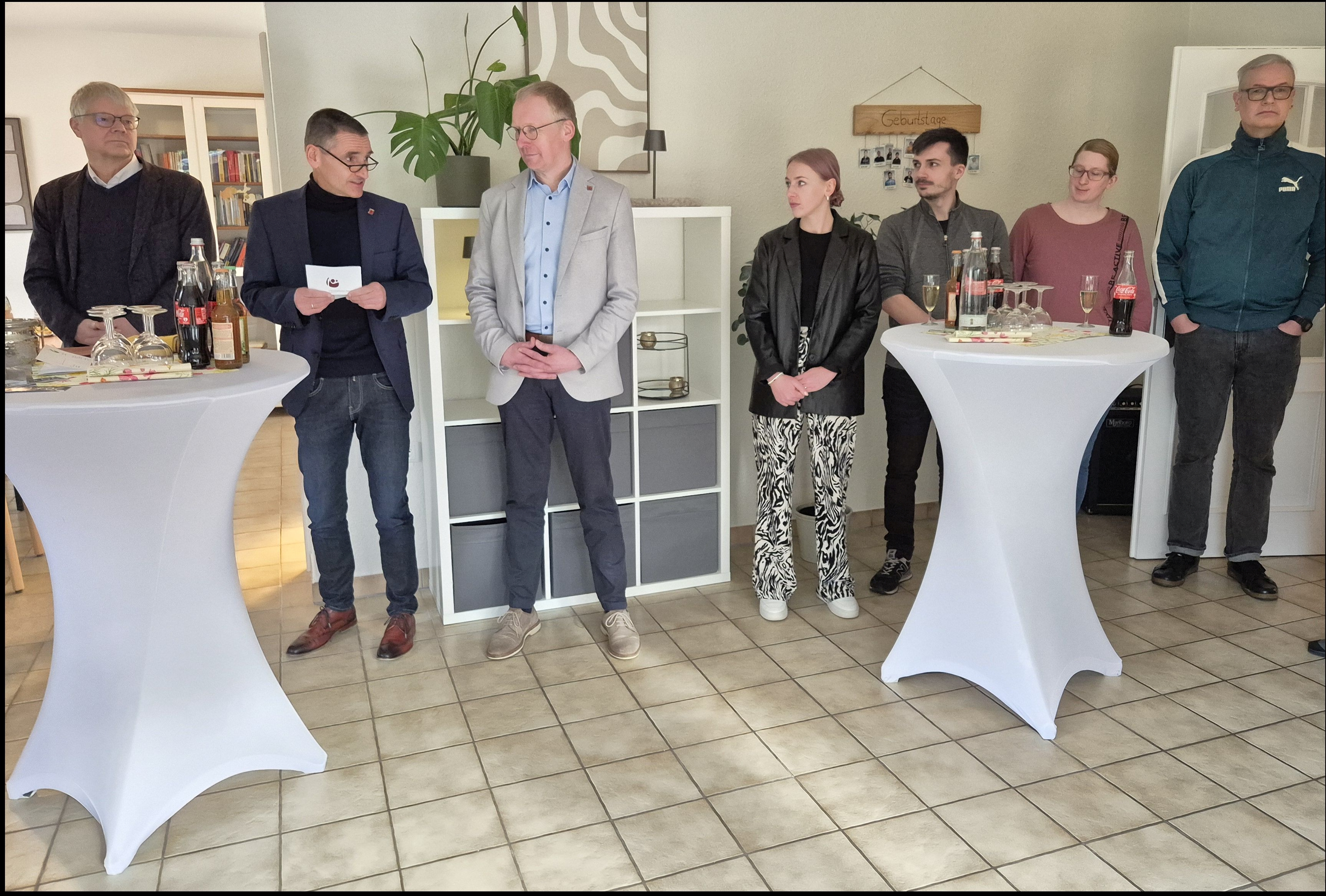 Mehrere Personen stehen bei der Einweihung der Wohngruppe Zedernweg in einem hellen Wohnraum nebeneinander. Zwei Stehtische mit Getr&auml;nken stehen im Vordergrund. Ein Mann spricht und h&auml;lt eine Karte in der Hand, w&auml;hrend die anderen aufmerksam zuh&ouml;ren.