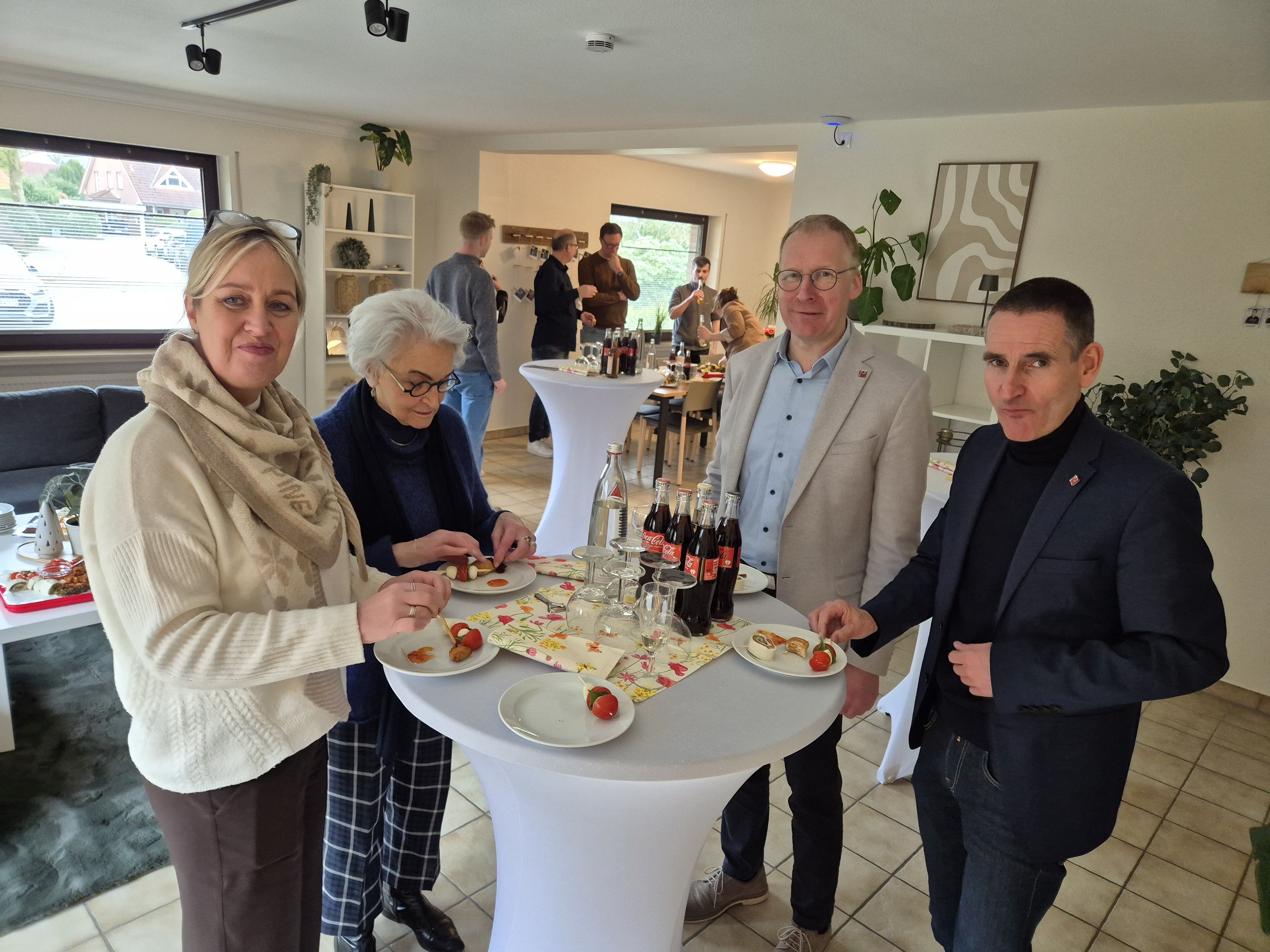 Vier G&auml;ste stehen bei der Einweihung der Wohngruppe Zedernweg an einem Stehtisch in einem hellen Wohnraum und essen kleine H&auml;ppchen. Auf dem Tisch stehen Getr&auml;nke und Gl&auml;ser. Im Hintergrund unterhalten sich weitere G&auml;ste in entspannter Atmosph&auml;re.