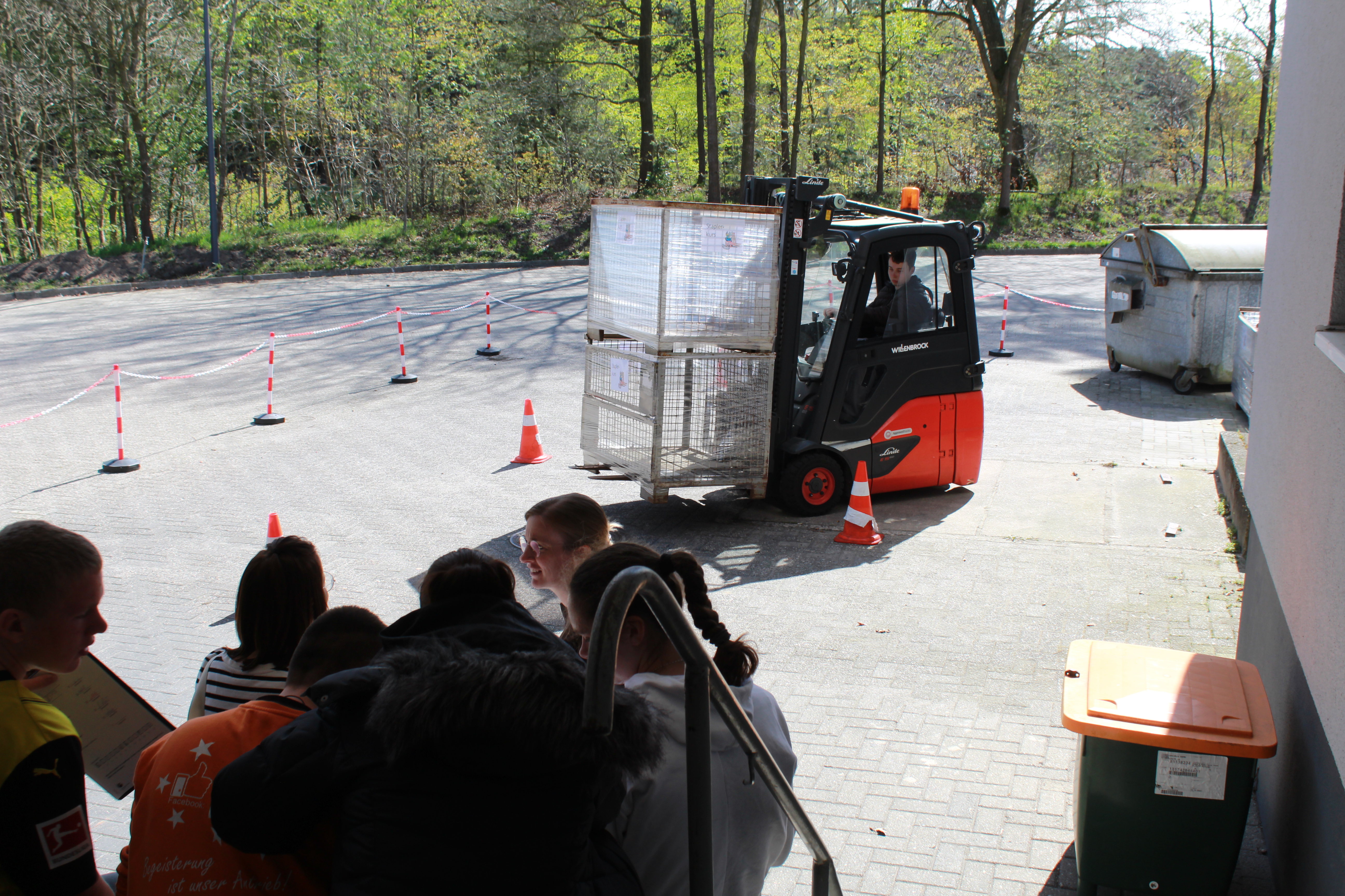 Ein Gabelstapler f&auml;hrt auf einem gepflasterten Hof Slalom um Pylonen, w&auml;hrend Jugendliche zuschauen.