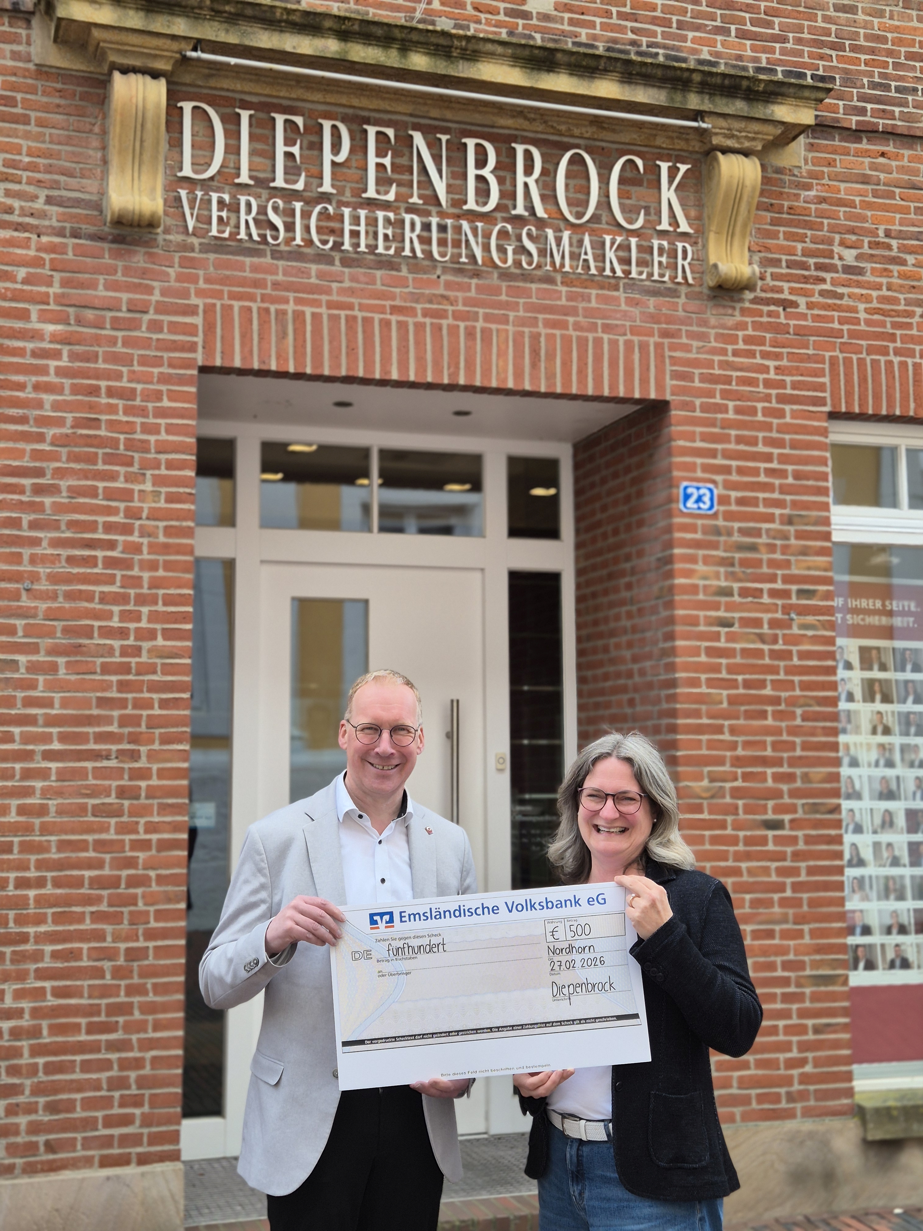 Ein Mann und eine Frau stehen bei gutem Wetter vor dem Geb&auml;ude der &bdquo;Diepenbrock Ver-sicherungsmakler GmbH&ldquo; und halten l&auml;chelnd einen Scheck &uuml;ber 500 Euro in der Hand.