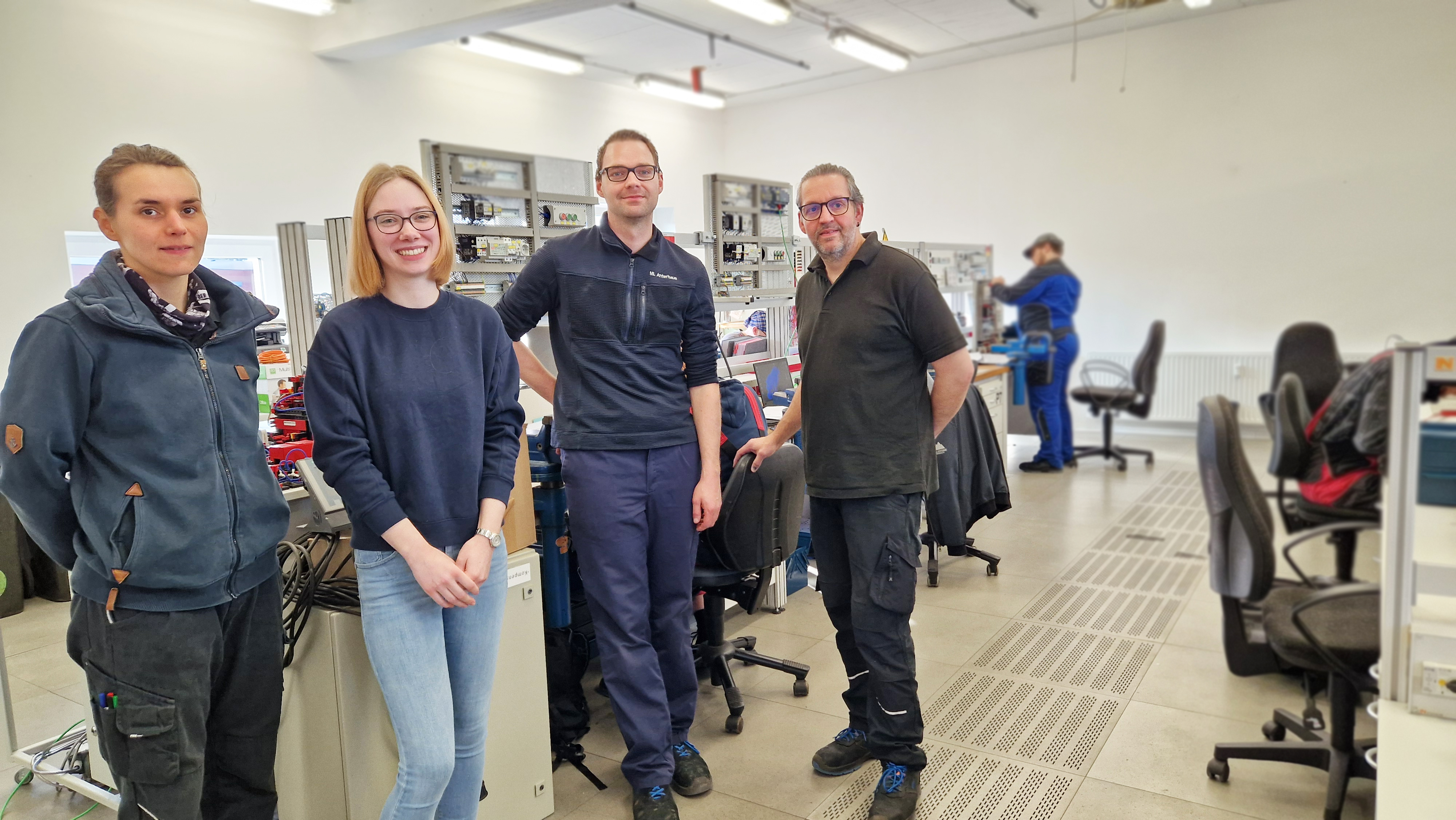 Auszubildende steht mit drei Ausbildern in einem Elektrotechnik-Schulungsraum, im Hintergrund Arbeitspl&auml;tze mit technischen Anlagen und weiteren Teilnehmenden.