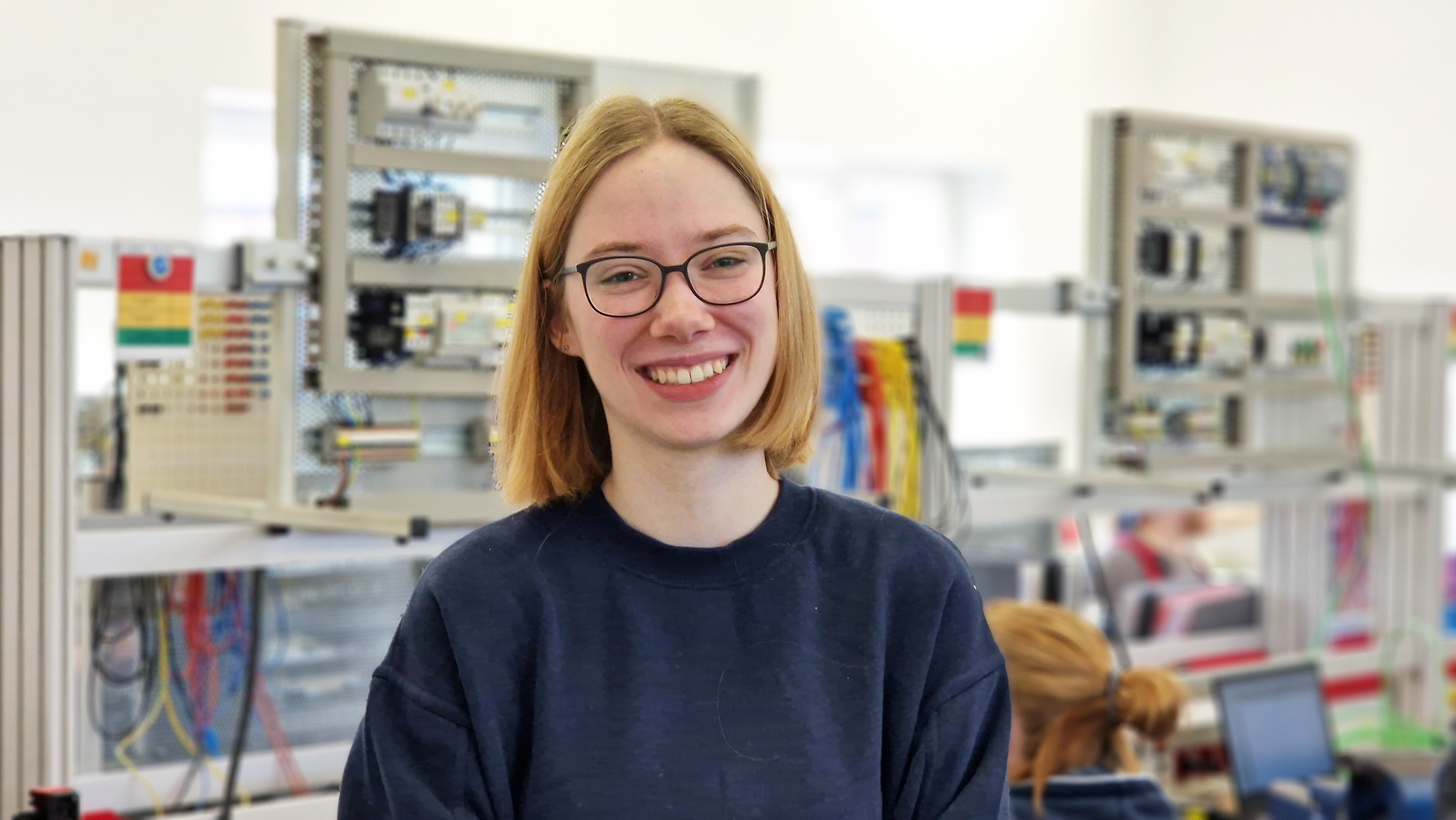 Junge Frau mit Brille l&auml;chelt in einem Elektrotechnik-Ausbildungsraum vor aufgebauten Schaltanlagen und Kabelmodulen.