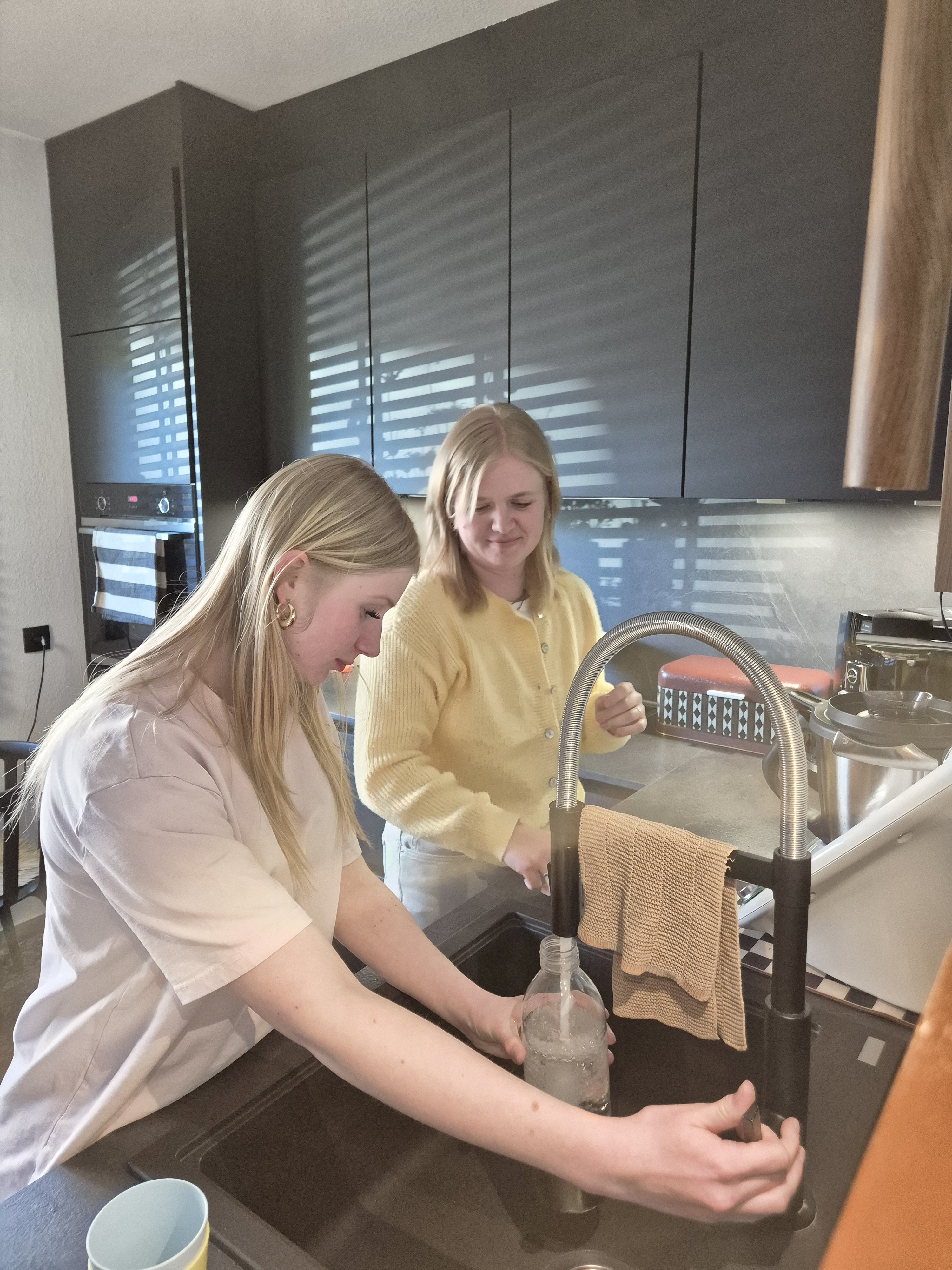 Eine junge Frau f&uuml;llt in einer modernen K&uuml;che eine Flasche mit Wasser am Sp&uuml;lbecken, w&auml;hrend eine zweite junge Frau danebensteht und sie dabei begleitet. Beide befinden sich an einer K&uuml;cheninsel mit dunklen Schr&auml;nken im Hintergrund und wirken konzentriert und entspannt im gemeinsamen Alltag.