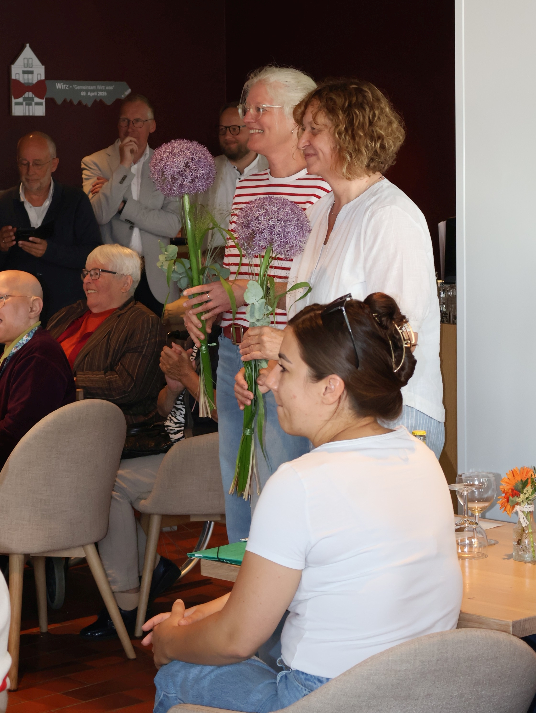 Zwei Frauen stehen in einem Veranstaltungsraum nebeneinander und halten jeweils einen Strau&szlig; mit gro&szlig;en violetten Zierlauchbl&uuml;ten in den H&auml;nden. Sie l&auml;cheln, w&auml;hrend die anwesenden G&auml;ste ihnen aufmerksam zuh&ouml;ren. Mehrere Personen sitzen an Tischen oder stehen im Hintergrund und verfolgen die Ehrung in einer freundlichen und wertsch&auml;tzenden Atmosph&auml;re.
