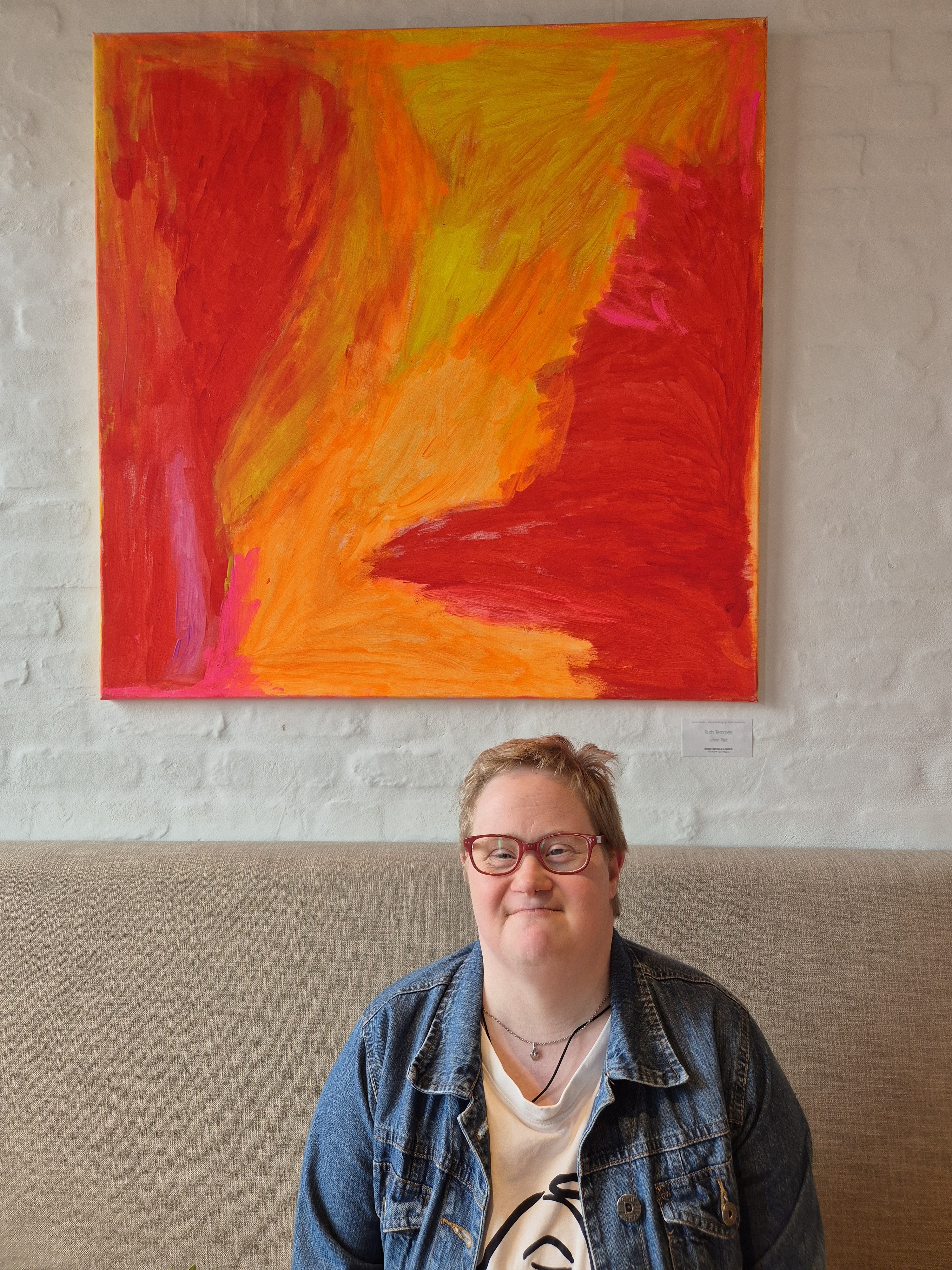Eine Frau mit kurzen Haaren und roter Brille sitzt vor einer hellen Wand auf einer gepolsterten Sitzbank. &Uuml;ber ihr h&auml;ngt ein gro&szlig;es abstraktes Gem&auml;lde in leuchtenden Rot-, Orange-, Gelb- und Pinkt&ouml;nen. Die Frau tr&auml;gt eine Jeansjacke &uuml;ber einem wei&szlig;en T-Shirt und blickt mit einem dezenten L&auml;cheln in die Kamera.
