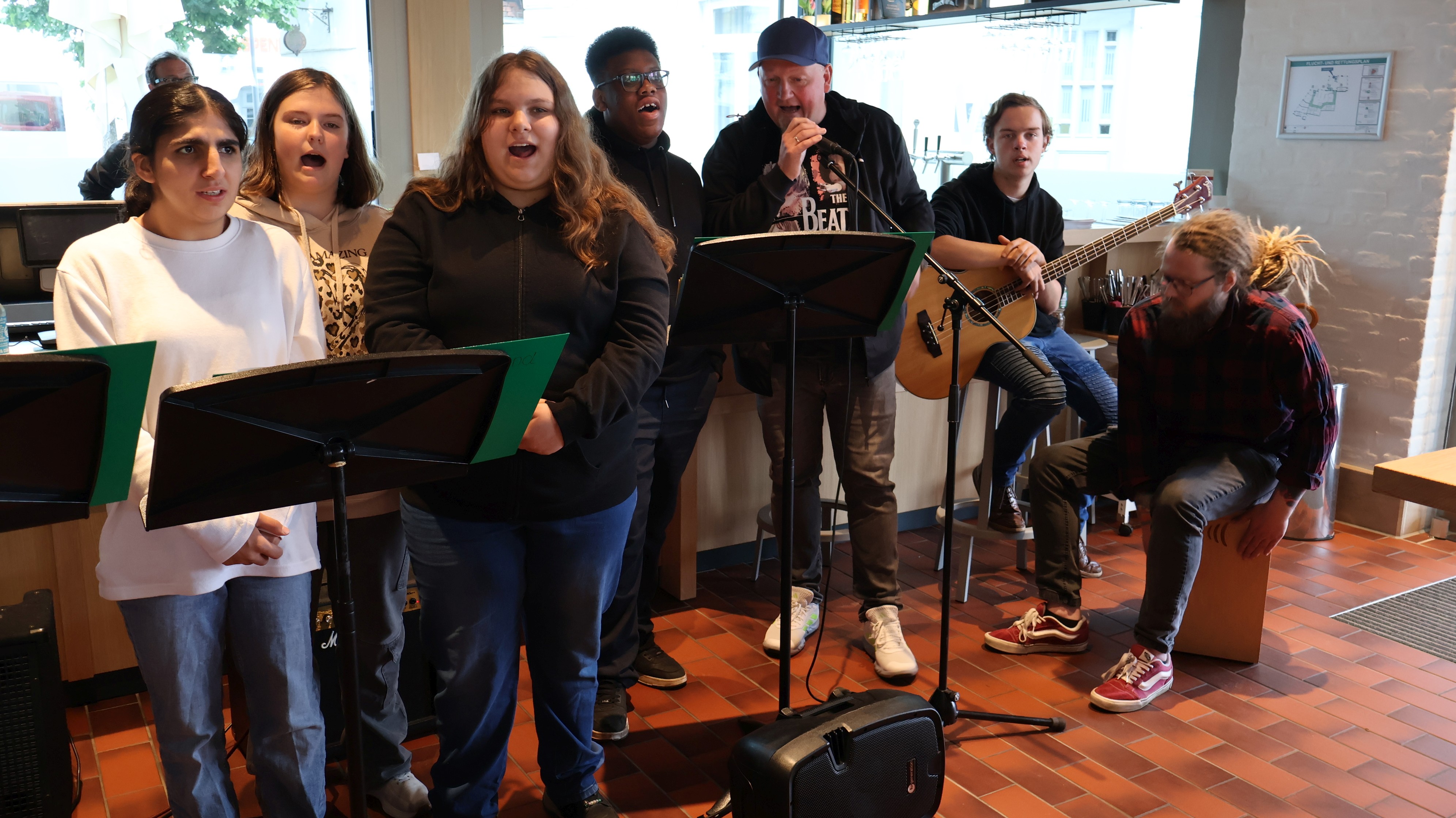 Mehrere Jugendliche und Erwachsene stehen gemeinsam in einem Caf&eacute; und musizieren. Einige singen hinter Notenst&auml;ndern, w&auml;hrend ein Musiker Gitarre spielt und ein weiterer auf einer Caj&oacute;n sitzt. Im Hintergrund sorgen gro&szlig;e Fenster f&uuml;r Tageslicht. Die Gruppe tritt konzentriert und mit sichtbarer Freude gemeinsam auf.