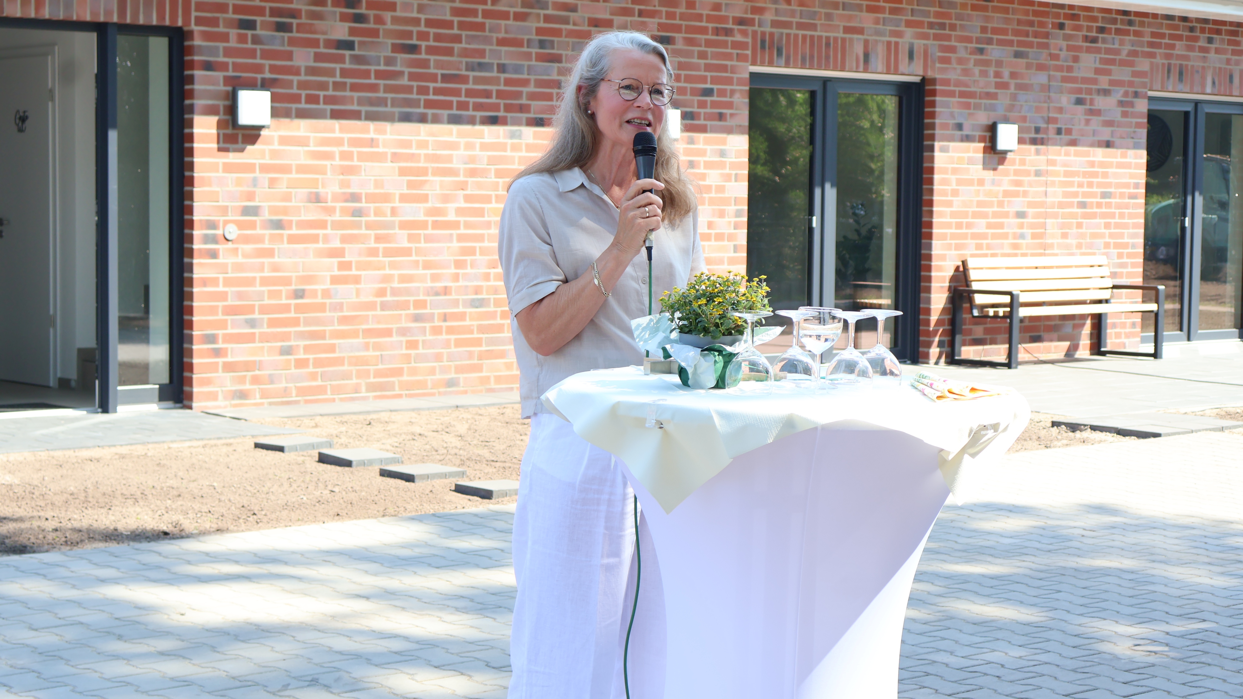 Klazina Hartholt, Bereichsleitung Kinder, Jugend und Familie im Christophorus-Werk, h&auml;lt bei der Er&ouml;ffnung der neuen Inobhutnahmewohngruppe in Klausheide eine Ansprache an einem Stehtisch mit Mikrofon. Im Hintergrund ist das neue Geb&auml;ude der Einrichtung bei sonnigem Wetter zu sehen.
