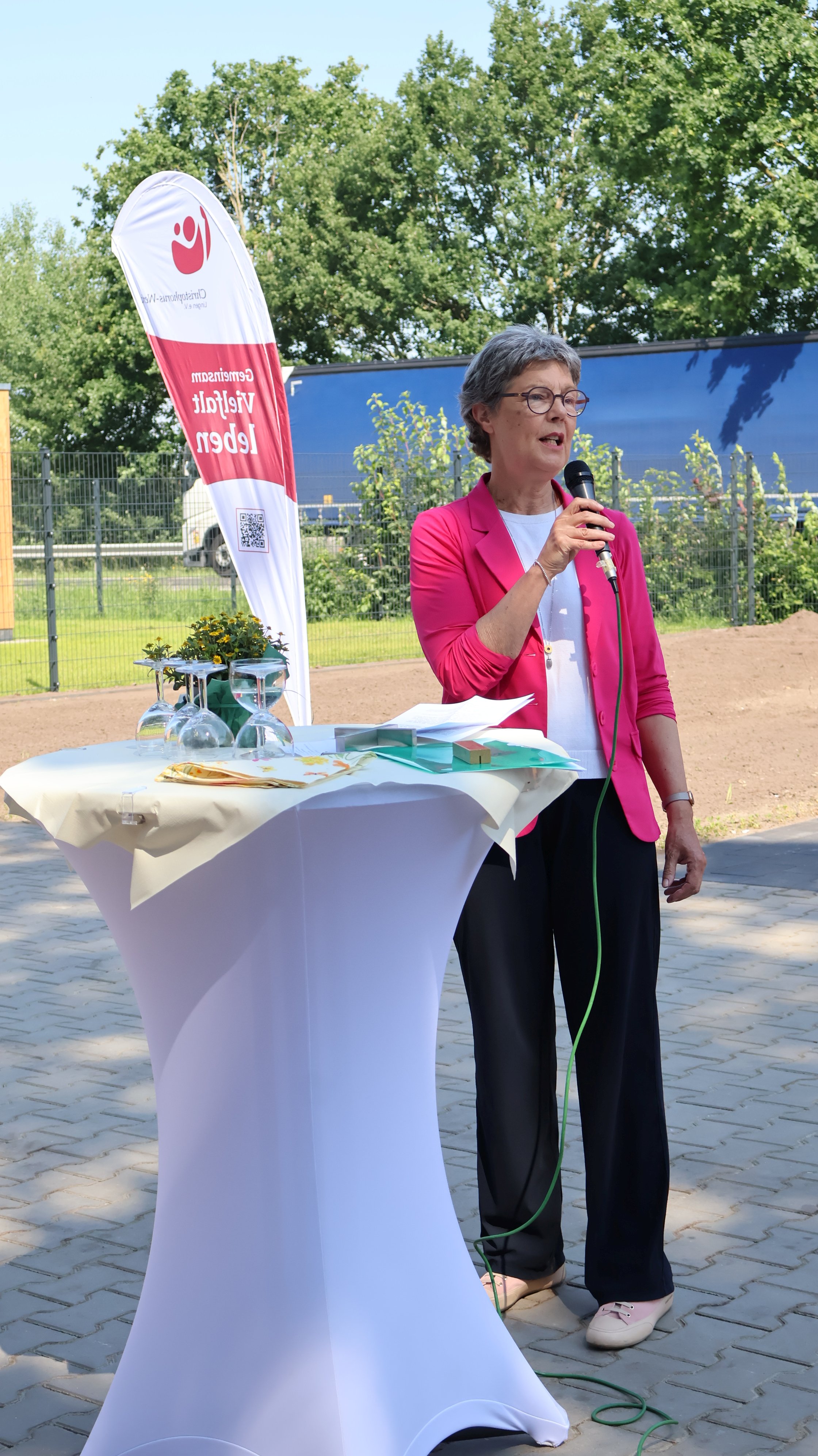 Gundula G&uuml;lker-Alsmeier, Kreisr&auml;tin des Landkreises Grafschaft Bentheim, h&auml;lt bei der Er&ouml;ffnung der neuen Inobhutnahmewohngruppe in Klausheide eine Ansprache. Sie spricht mit einem Mikrofon an einem Stehtisch im Au&szlig;enbereich der Einrichtung. Im Hintergrund sind ein Banner des Christophorus-Werks mit dem Claim &bdquo;Gemeinsam Vielfalt leben&ldquo; sowie B&auml;ume zu sehen.
