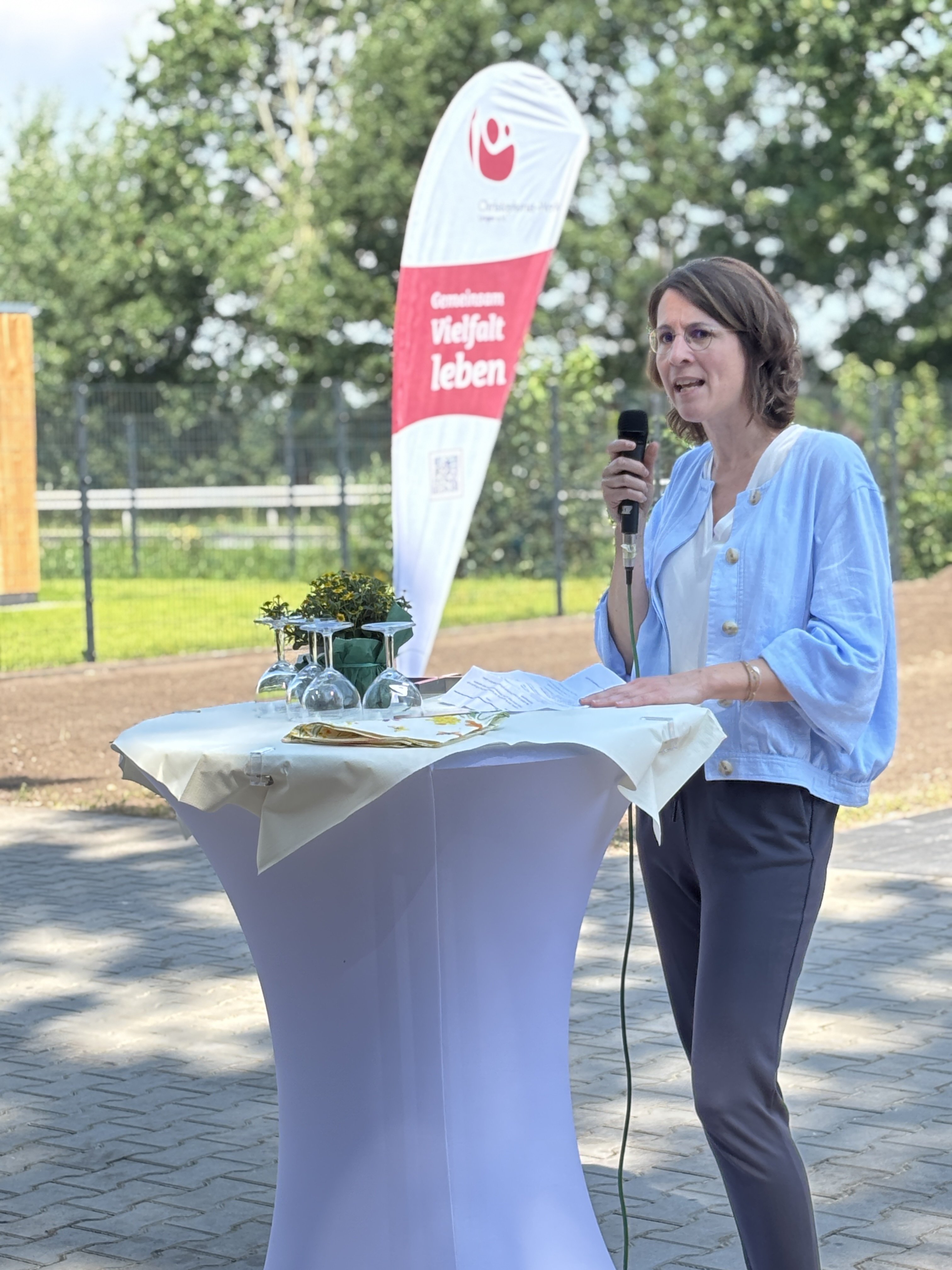 Katrin M&ouml;llenkamp, Stadtr&auml;tin der Stadt Lingen, h&auml;lt bei der Er&ouml;ffnung der neuen Inobhutnahmewohngruppe in Klausheide eine Ansprache. Sie steht mit einem Mikrofon an einem Stehtisch im Au&szlig;enbereich der Einrichtung. Im Hintergrund sind ein Banner des Christophorus-Werks mit dem Claim &bdquo;Gemeinsam Vielfalt leben&ldquo; sowie B&auml;ume zu sehen.