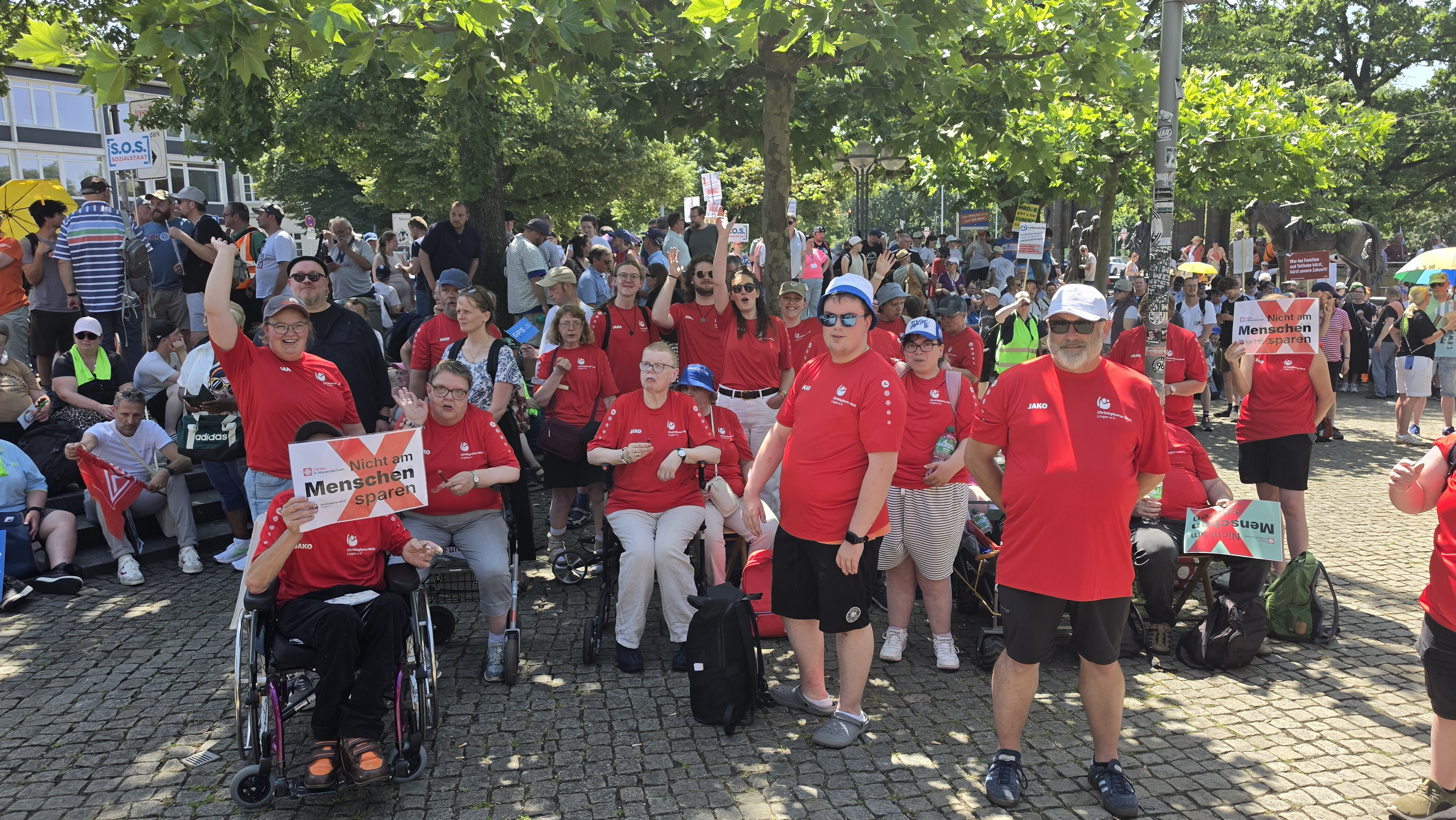 Gruppe Menschen mit roten Shirts auf einem Platz. Im Hintergrund stehen zahlreiche weitere Menschen mit Plakaten und Fahnen. 