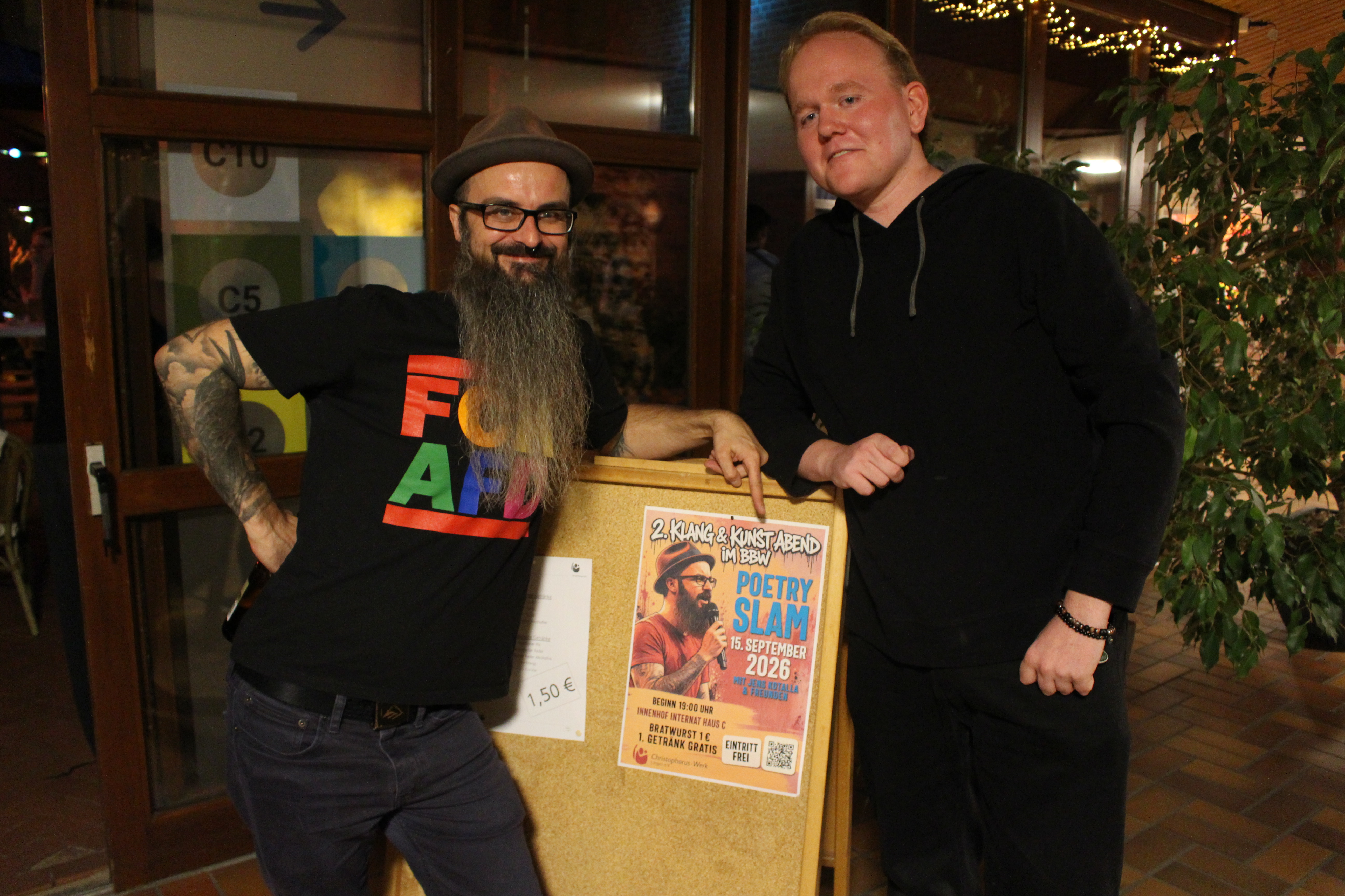 Die Comedians Jens Kotalla und Julian Deters stehen nach dem &bdquo;Klang & Kunst&ldquo;-Abend im BBW Lingen nebeneinander. Zwischen ihnen befindet sich ein Aufsteller mit dem Veranstaltungsplakat.