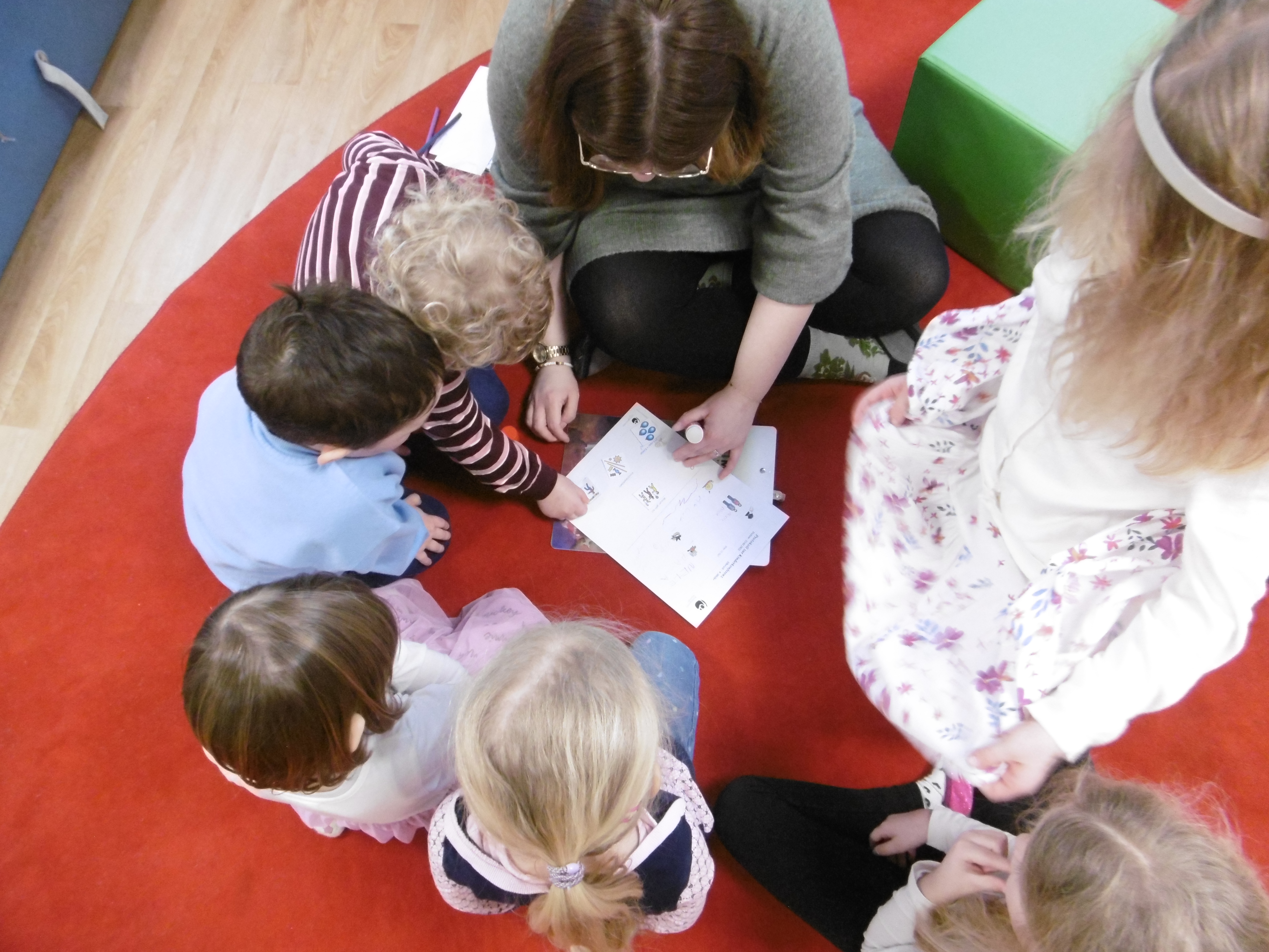 Eine Erzieherin sitzt mit einer Gruppe Kinder im Kreis auf dem Boden. Gemeinsam schauen sie auf ein Blatt Papier und diskutieren. 