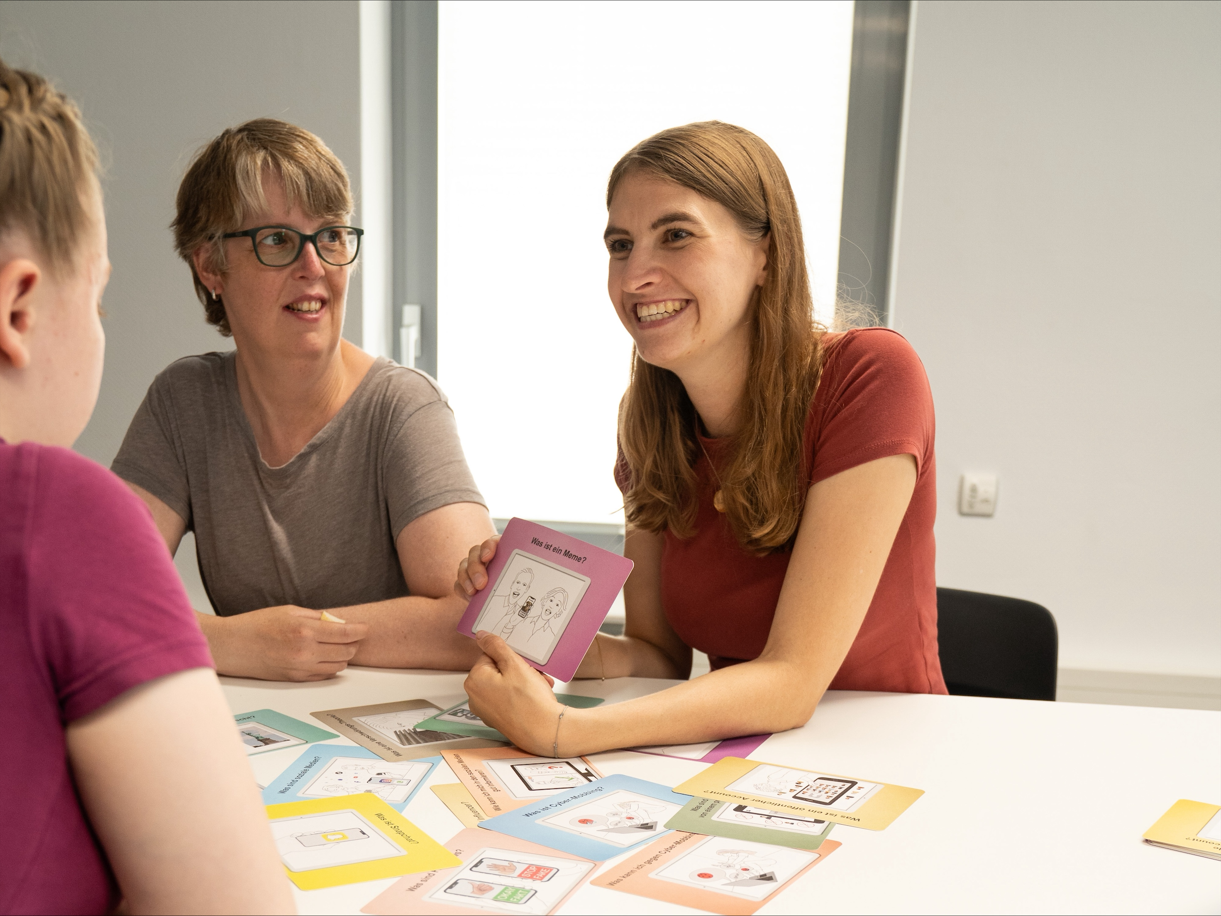 Drei Frauen sitzen am Tisch. Eine Frau zeigt den anderen eine Karte.
