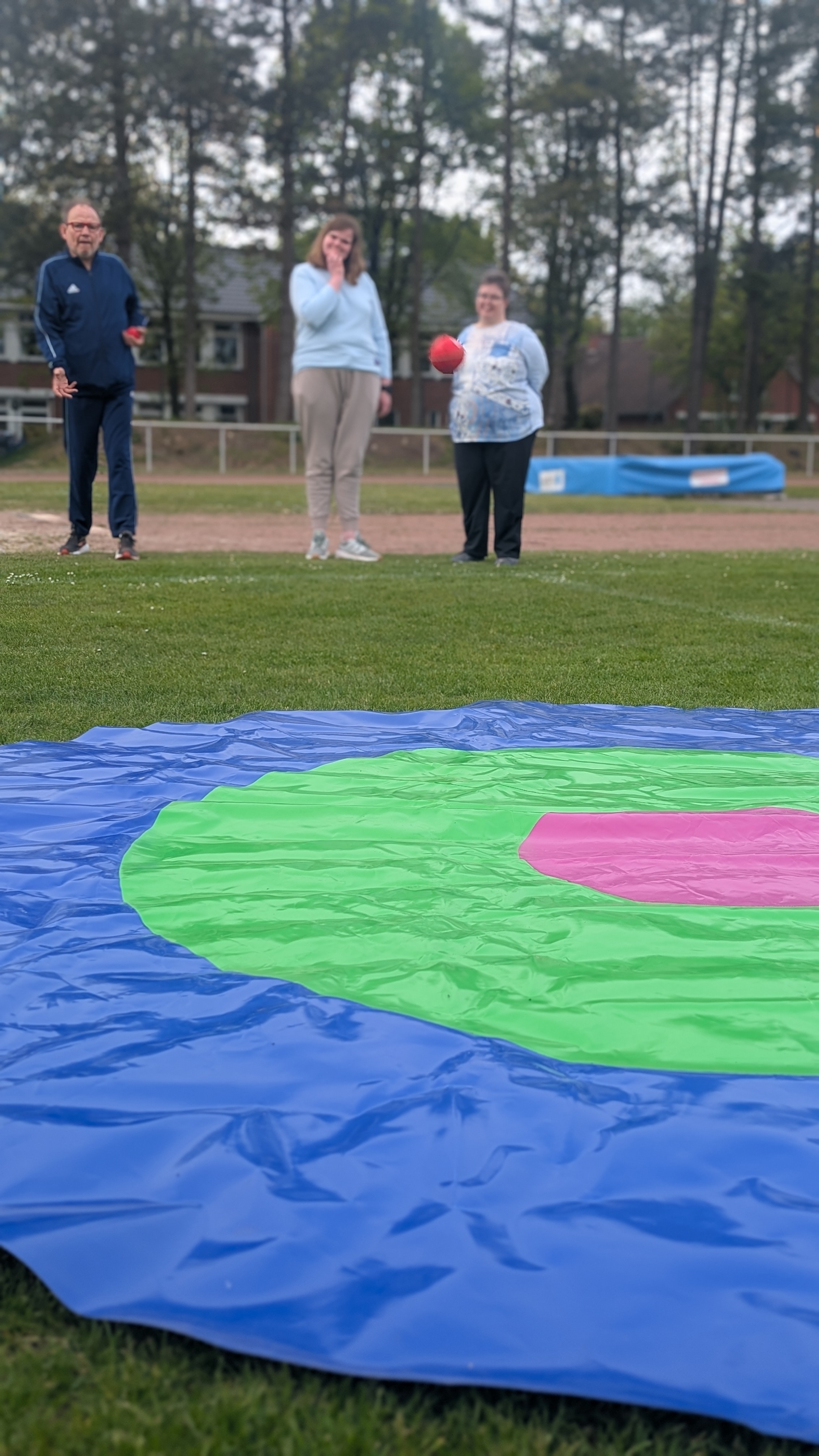 Eine gro&szlig;e Zielscheibe liegt auf dem Gras und ein Mann wirft dahin B&auml;lle. Zwei Frauen stehen daneben.
