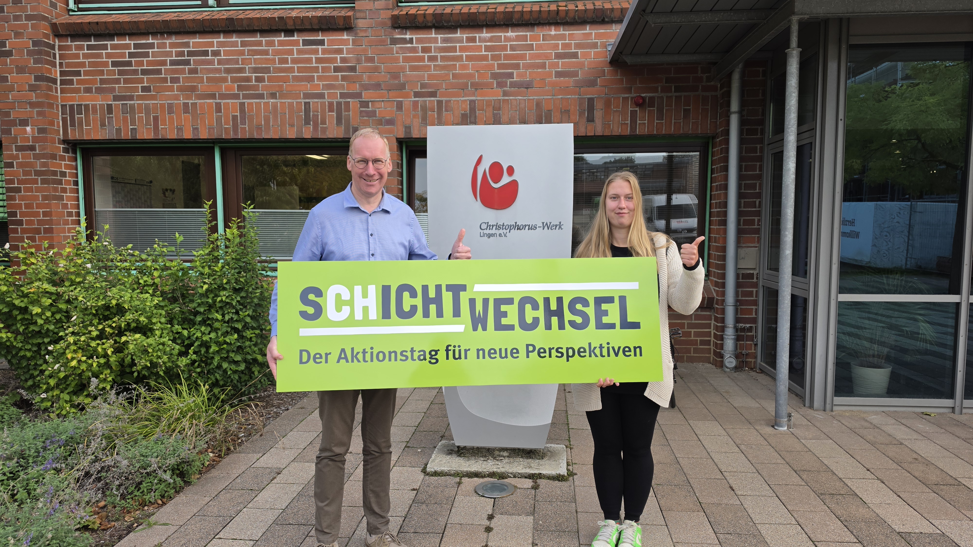 Gesch&auml;ftsf&uuml;hrer Stefan Kerk und Werkstattbesch&auml;ftigte Josephine stehen vor dem Christophorus-Werk in Lingen und halten gemeinsam ein gr&uuml;nes Schild mit der Aufschrift &bdquo;Schichtwechsel &ndash; Der Aktionstag f&uuml;r neue Perspektiven&ldquo;. Beide zeigen mit dem Daumen nach oben.