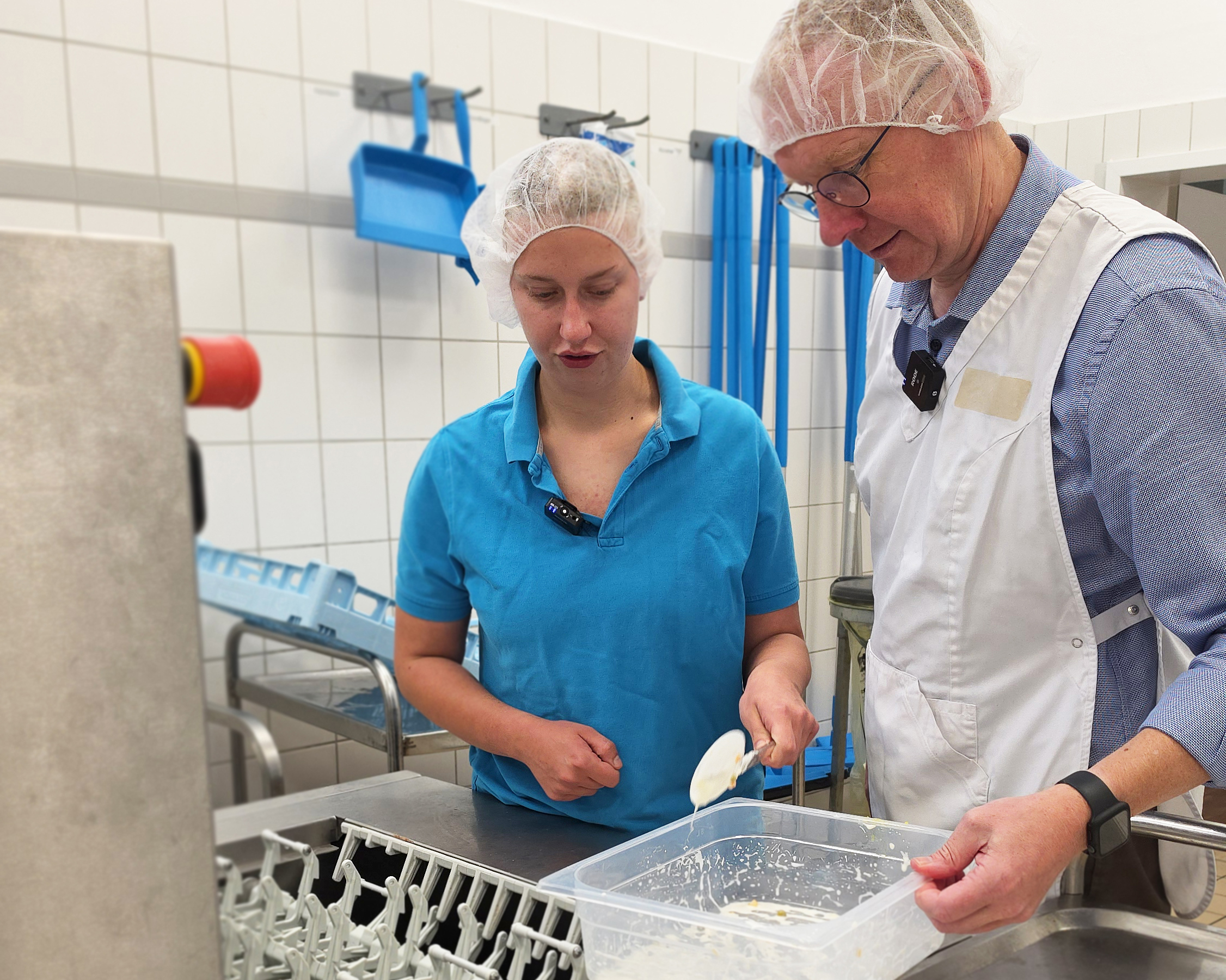 Werkstattbesch&auml;ftigte Josephine und Gesch&auml;ftsf&uuml;hrer Stefan Kerk stehen gemeinsam in der K&uuml;che der WfbM an einer Sp&uuml;lmaschine. Beide tragen Haarnetze. Josephine zeigt Stefan einen Arbeitsschritt beim Einr&auml;umen beziehungsweise Reinigen des Geschirrs.