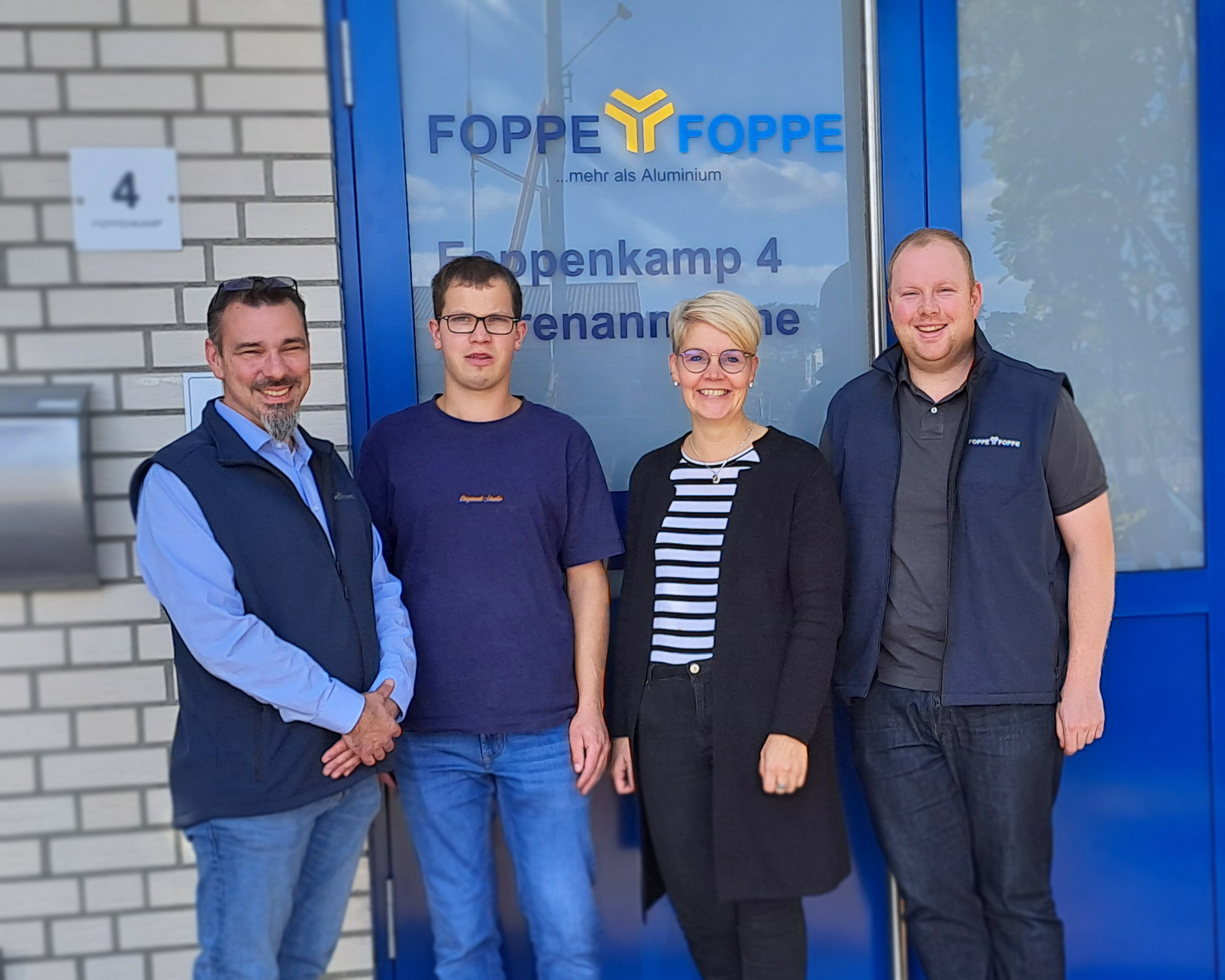 Werkstattbesch&auml;ftigter Christian Hinken steht gemeinsam mit drei Mitarbeitenden von Foppe & Foppe vor dem Firmengeb&auml;ude in Lengerich. Im Hintergrund sind der Schriftzug und das Logo von Foppe & Foppe zu sehen.