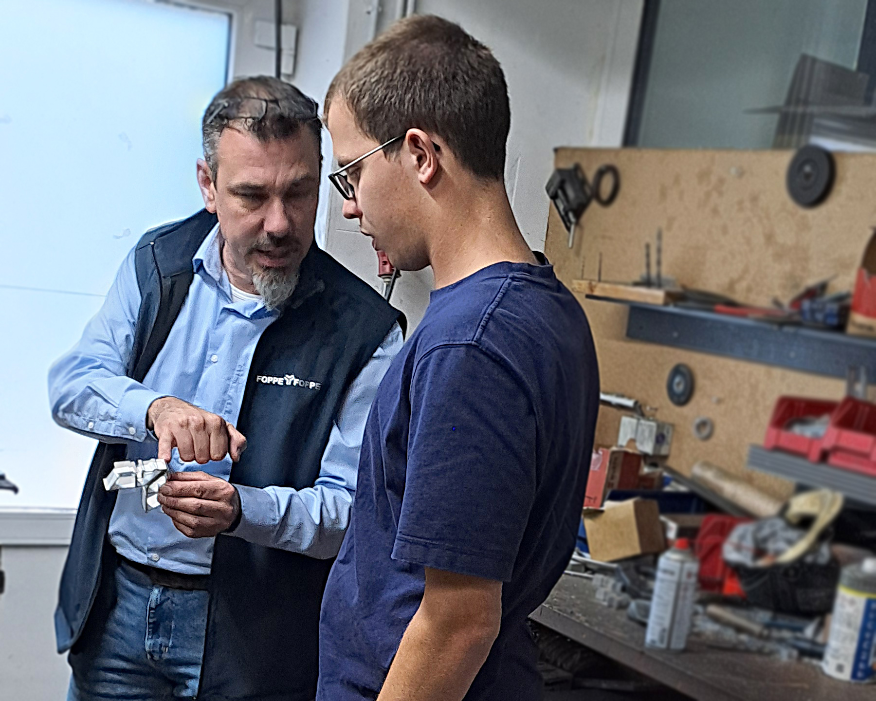 Werkstattbesch&auml;ftigter Christian Hinken steht gemeinsam mit einem Mitarbeiter von Foppe & Foppe in der Metallwerkstatt. Der Mitarbeiter zeigt ihm ein Bauteil aus Aluminium und erkl&auml;rt einen Arbeitsschritt.