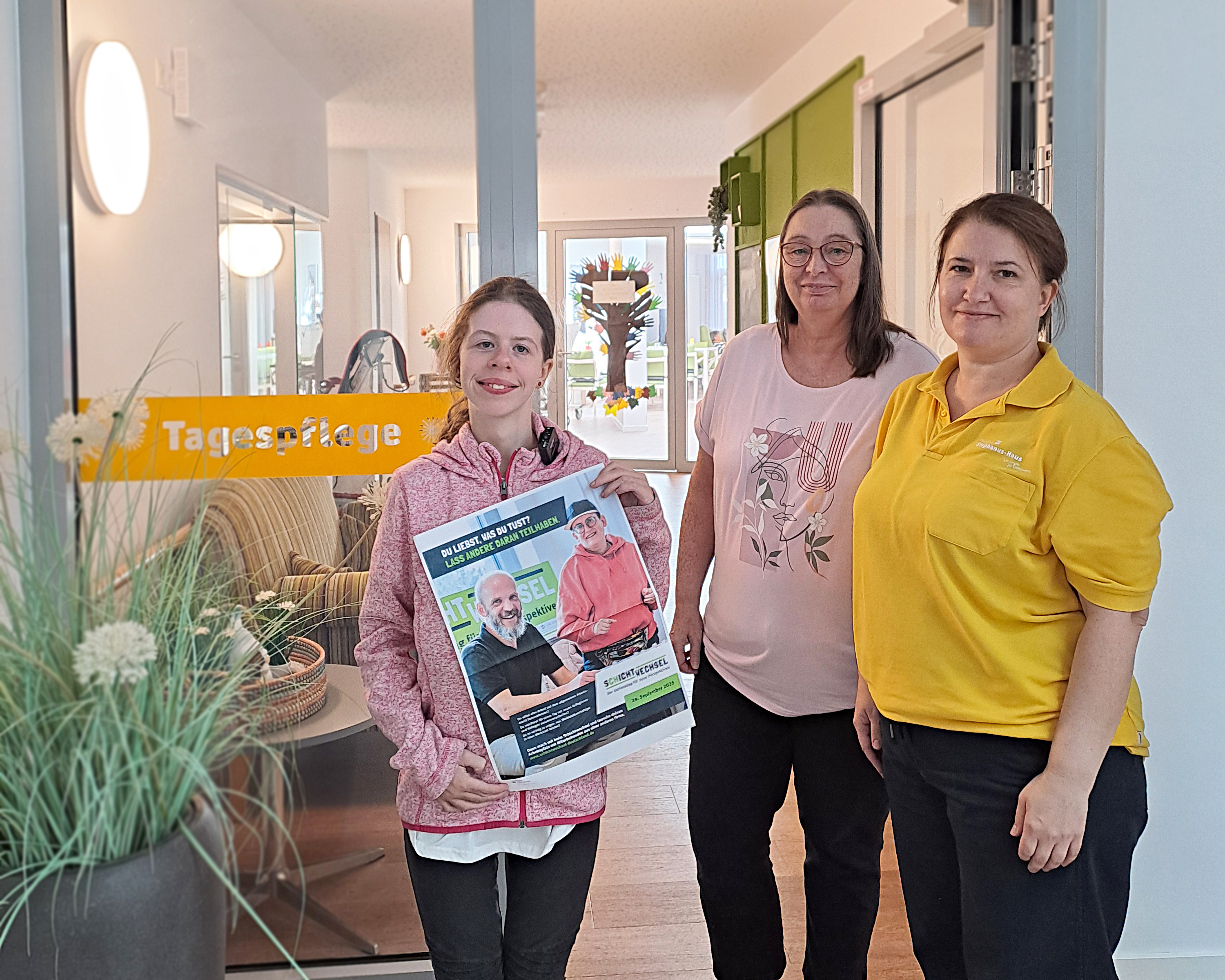 Werkstattbesch&auml;ftigte Lilli steht gemeinsam mit zwei Mitarbeiterinnen in der Tagespflege des Stephanus-Hauses. Lilli h&auml;lt ein Plakat zum Aktionstag &bdquo;Schichtwechsel&ldquo; in den H&auml;nden. Im Hintergrund ist der Eingangsbereich der Tagespflege zu sehen.