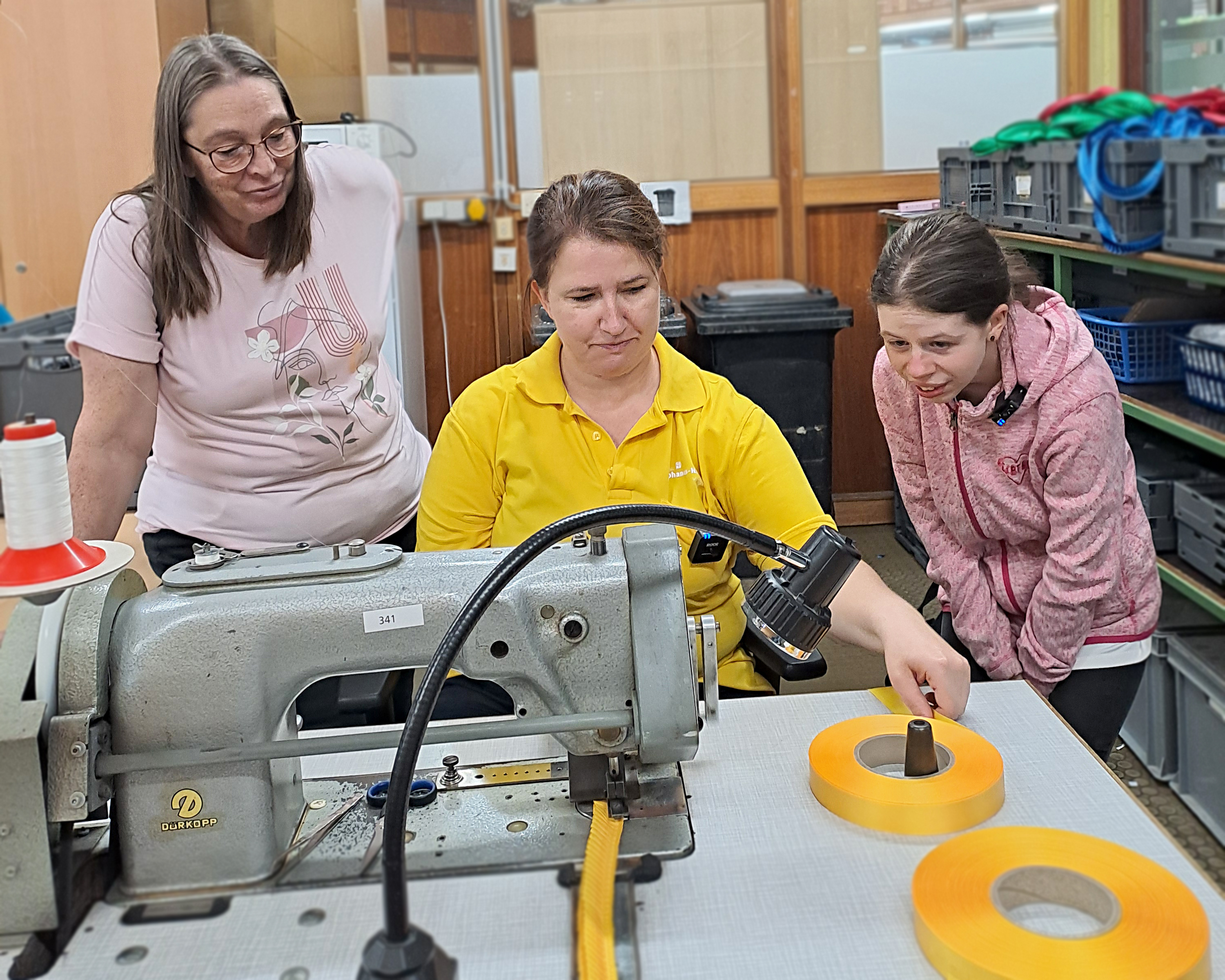 Lilli und zwei Mitarbeiterinnen stehen an einer N&auml;hmaschine in der Schleifenwerkstatt des Christophorus-Werkes. Eine Mitarbeiterin sitzt an der Maschine und zeigt einen Arbeitsschritt mit gelbem Band, w&auml;hrend Lilli und die zweite Mitarbeiterin aufmerksam zusehen.