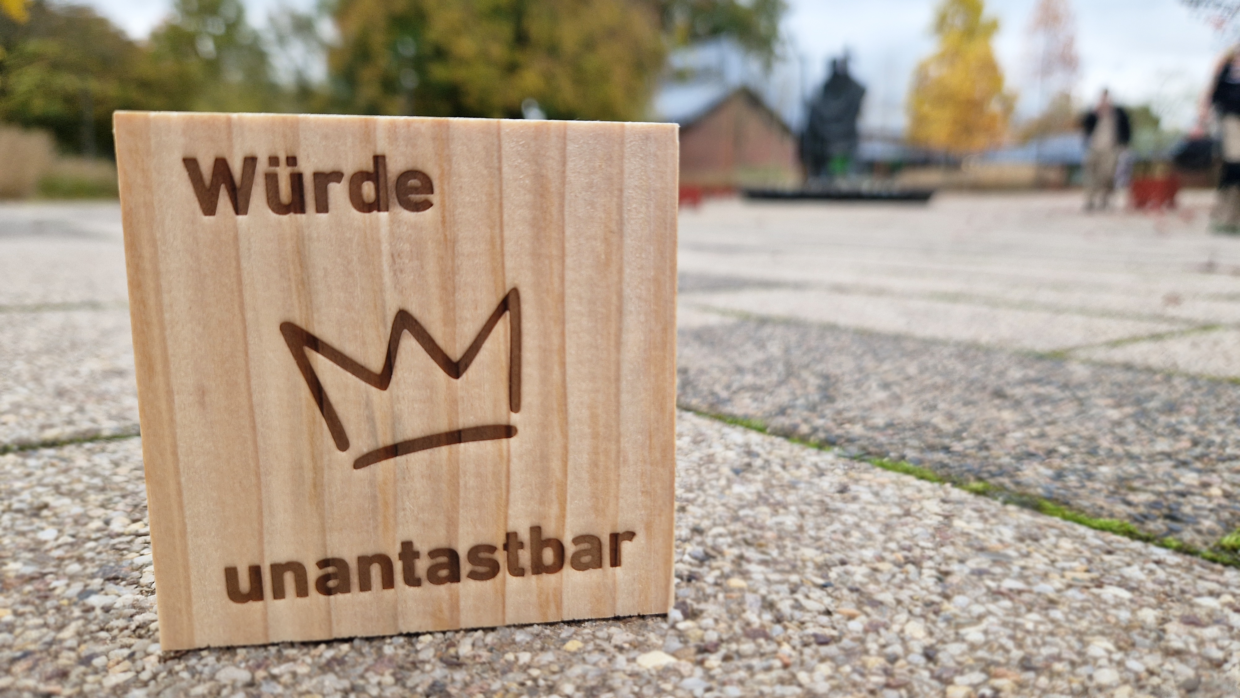 Ein Holzstück steht auf einem Platz vor einem Brunnen mit einer Statue.. Auf dem Holz wurde das Bild einer Krone eingebrannt und die Wörter: „Würde unantastbar“