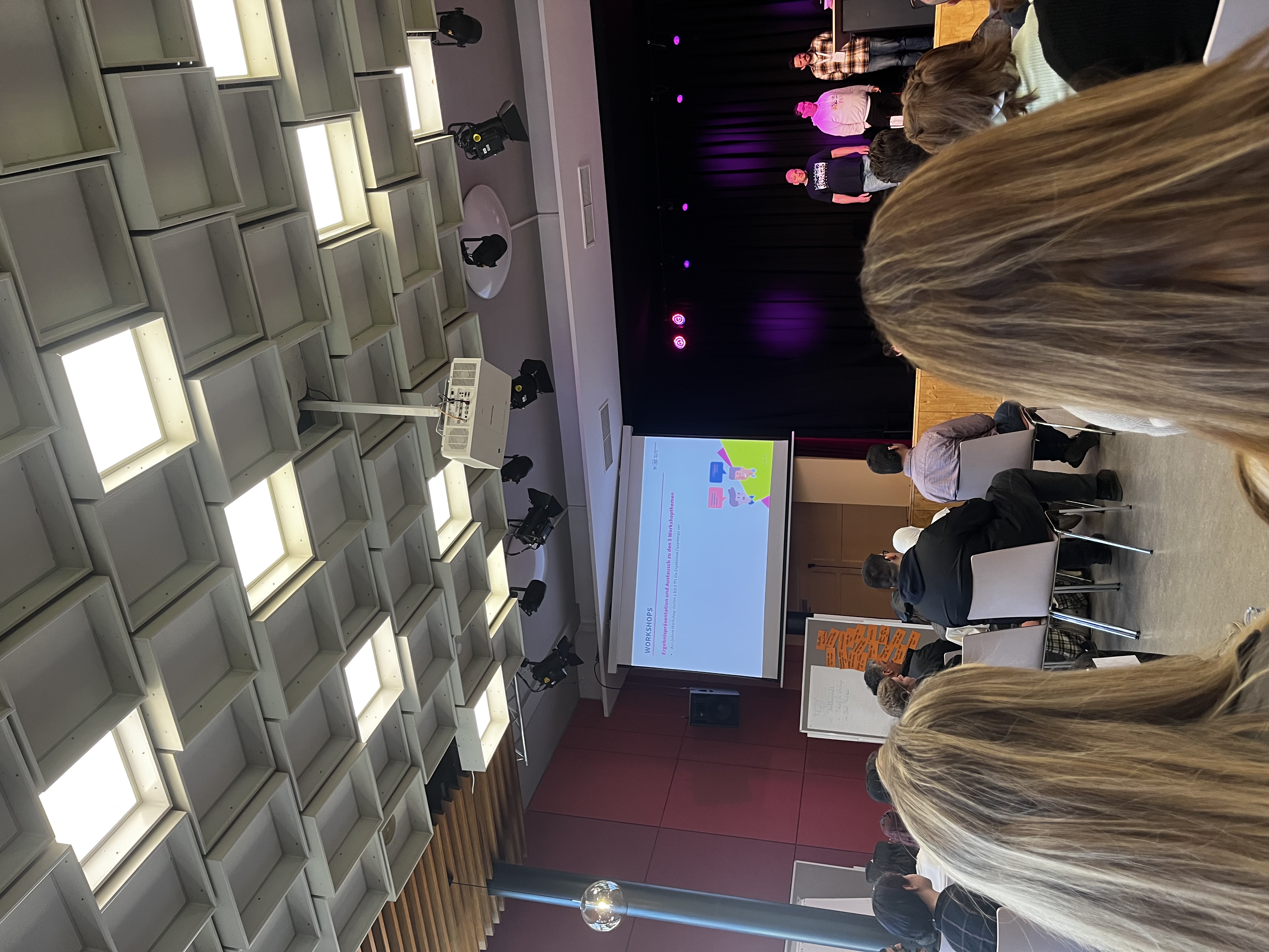 Das Foto zeigt einen Workshopraum mit sitzenden Teilnehmenden, einer B&uuml;hne und Pr&auml;sen-tationsfolie. Drei Personen stehen vorne auf der B&uuml;hne und sprechen zum Publikum.