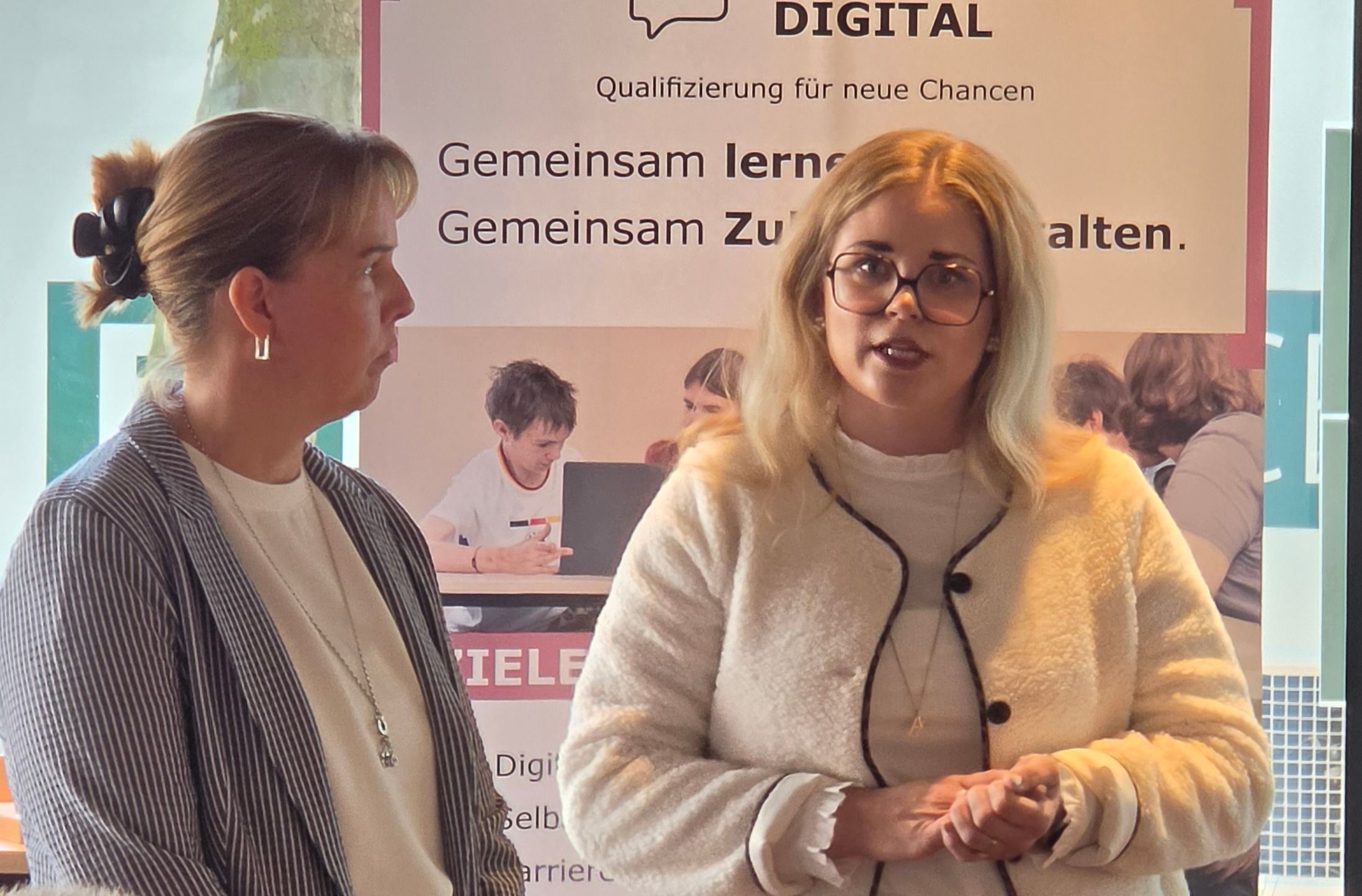 Zwei Projektleiterinnen stellen das Projekt KompetenzSprung vor. Im Hintergrund befindet sich Pr&auml;sentationsmaterial.