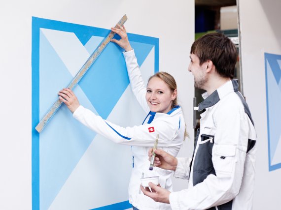 Anleiterin und Auszubildender malen ein blaues Muster auf eine weiße Wand.