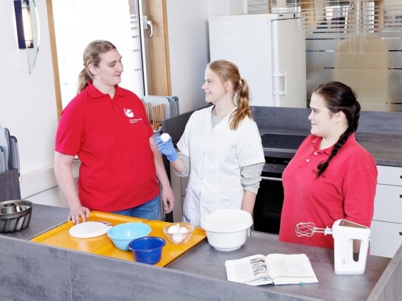 Anleiterin zeigt zwei Auszubildenden die Zutaten für das Backen.