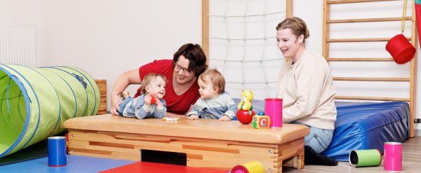 Mitarbeiterin, Mutter und zwei Säuglinge sitzen auf dem Boden einer Turnhalle.